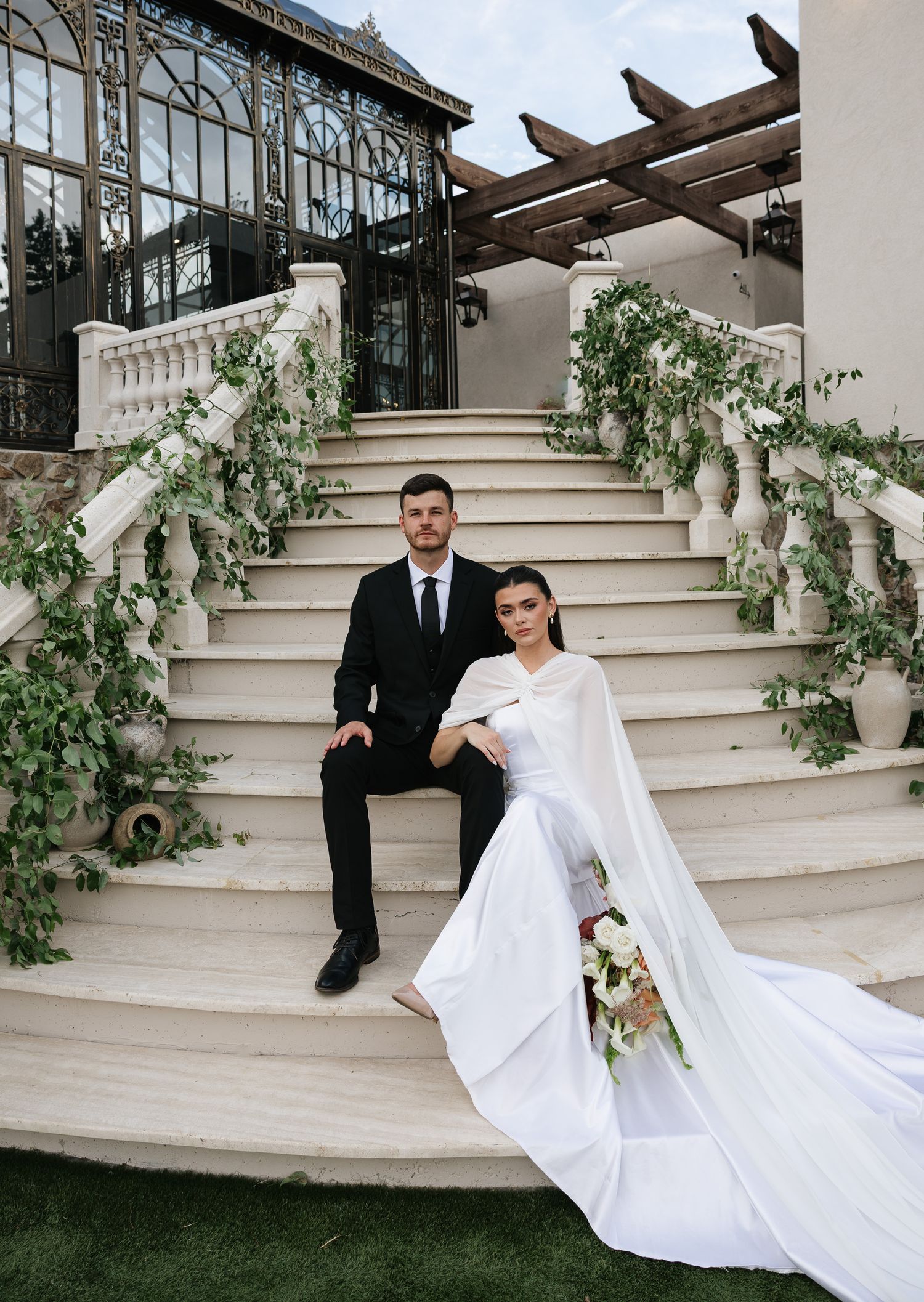 Bride and groom posing on elegant white stone staircase adorned with greenery outside a grand villa.