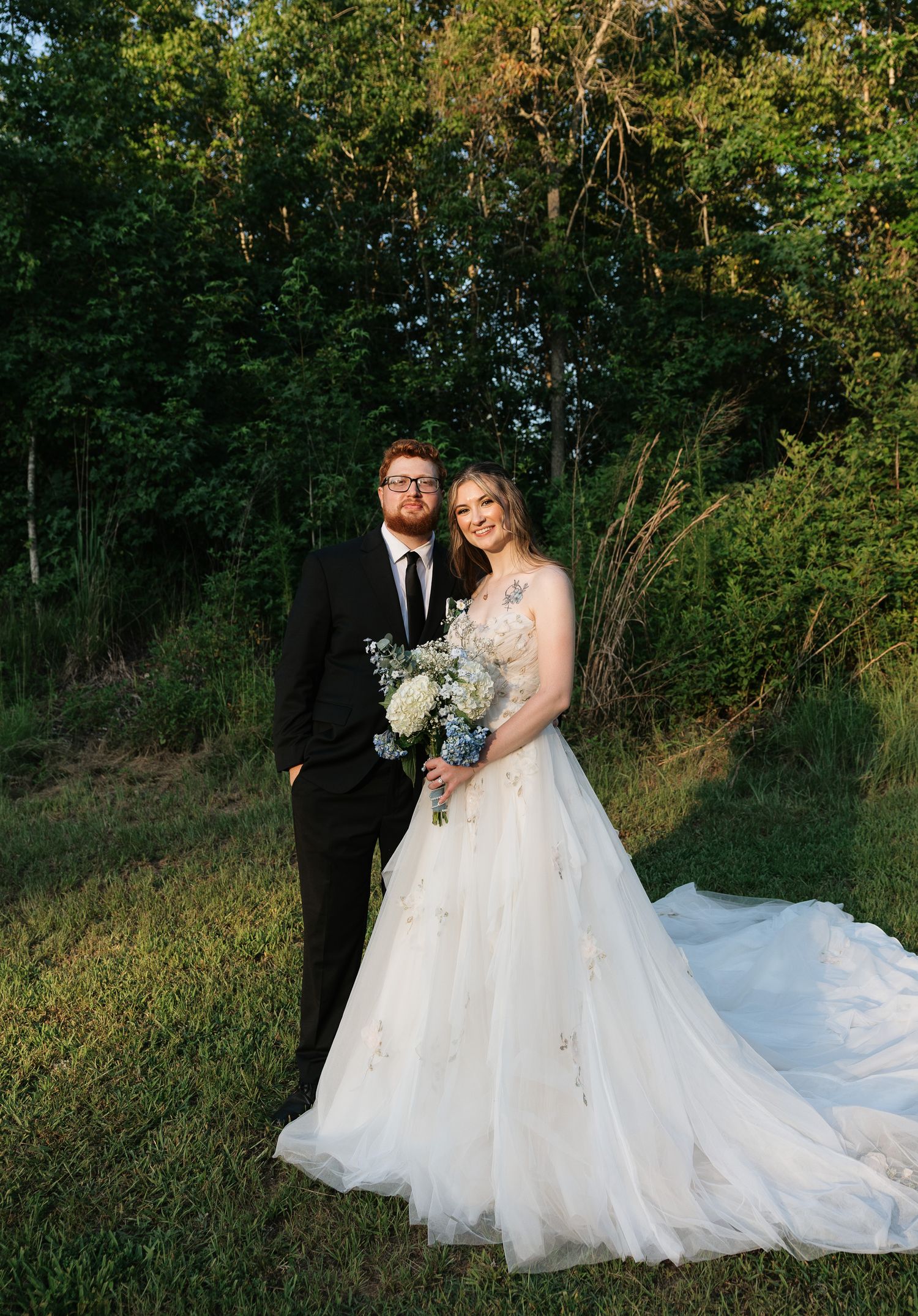 Bride in floral ballgown holds bouquet beside groom in black suit, posing outdoors near lush green trees.