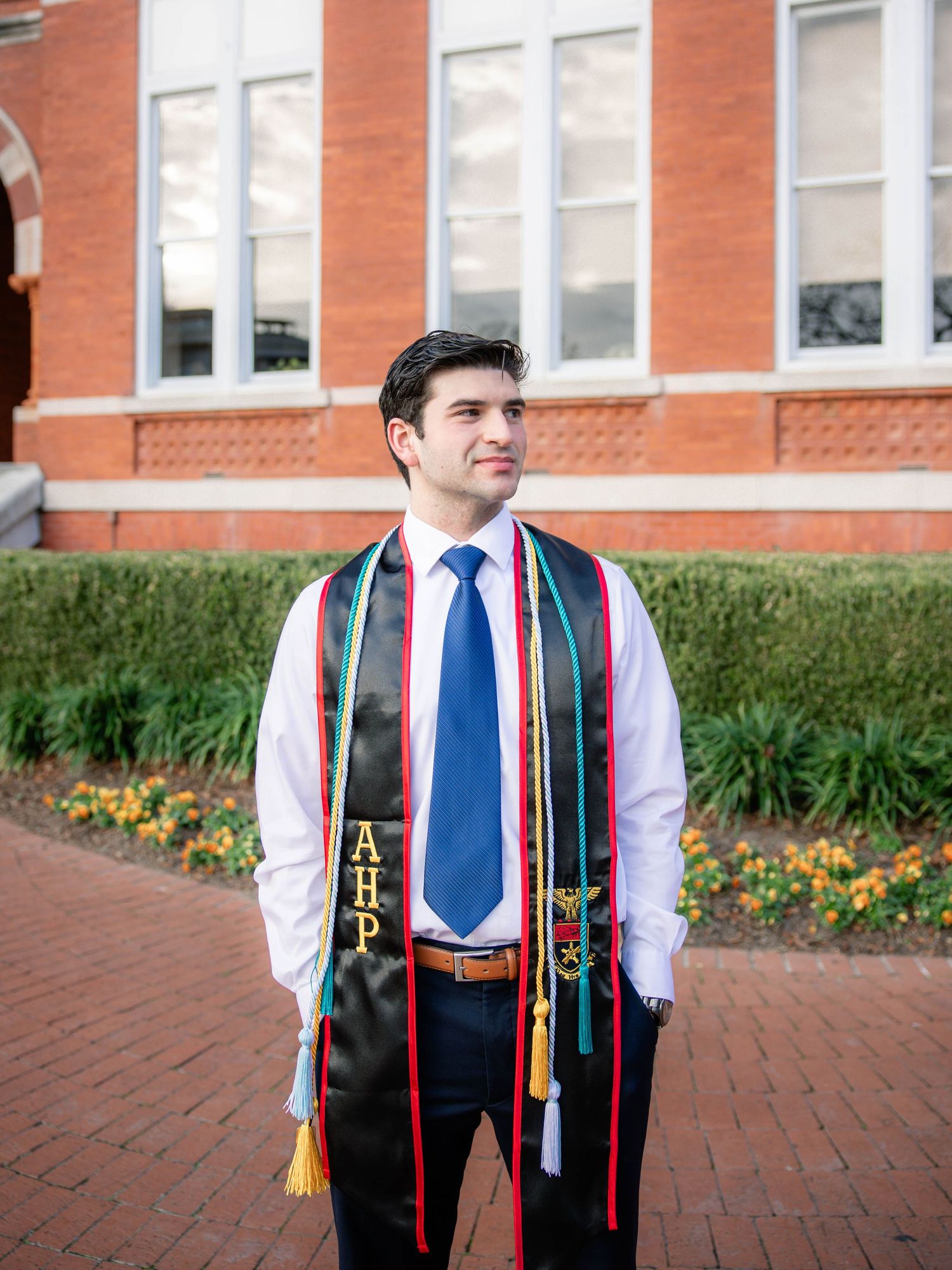 Graduate wearing white dress shirt, blue tie and black vest with colorful stole poses on brick walkway near building.