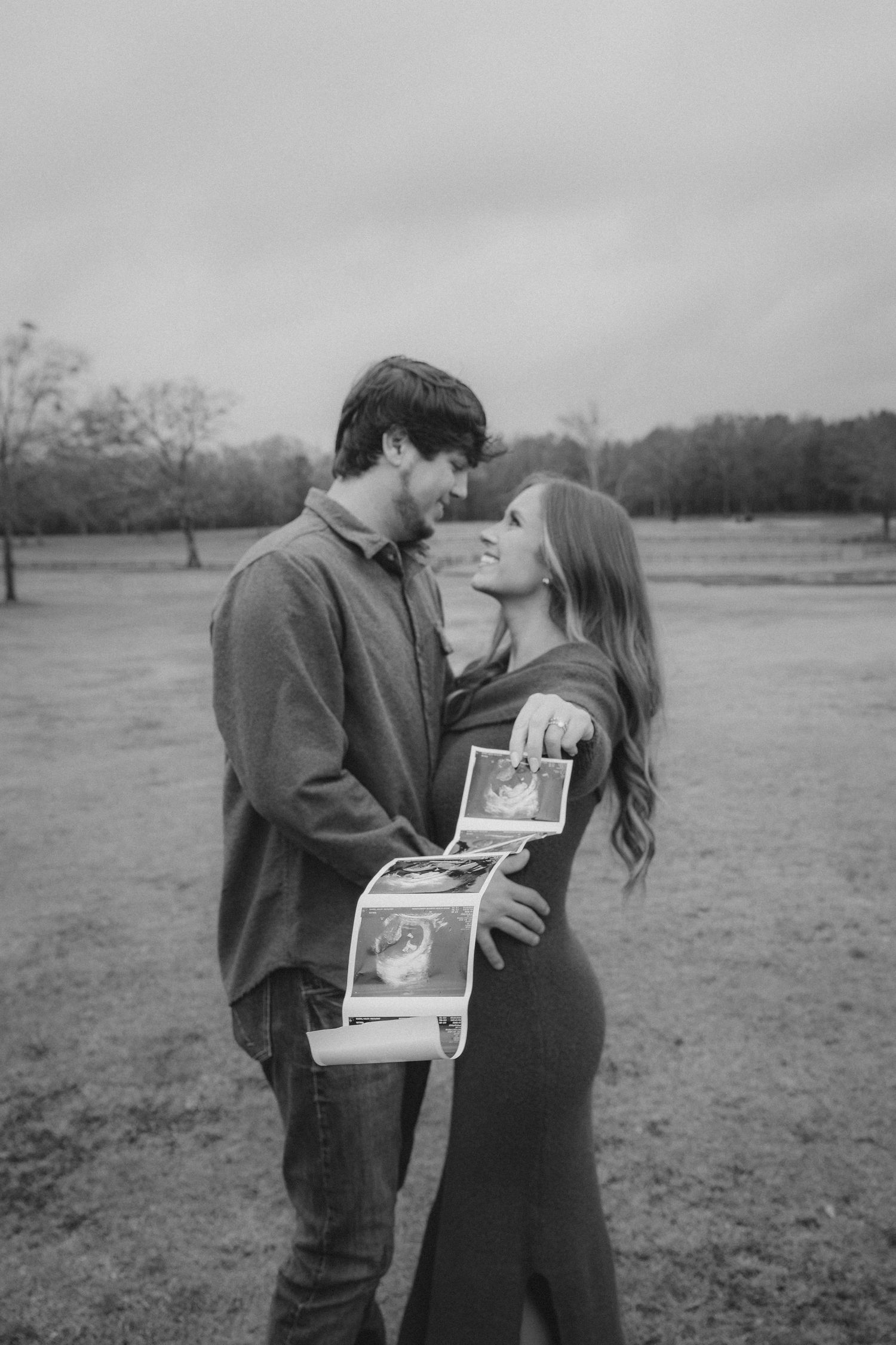 Black and white photo of a couple smiling at each other outdoors, holding ultrasound photos for a pregnancy announcement.