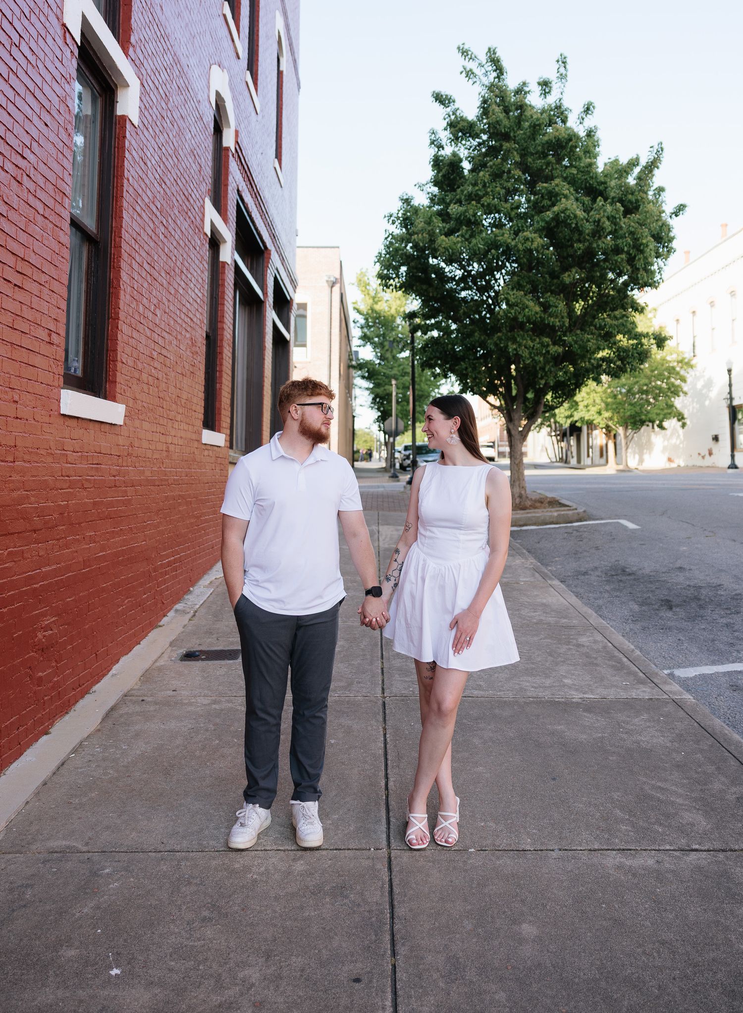 A young couple in white outfits holds hands on a city sidewalk beside a red brick building and green tree.