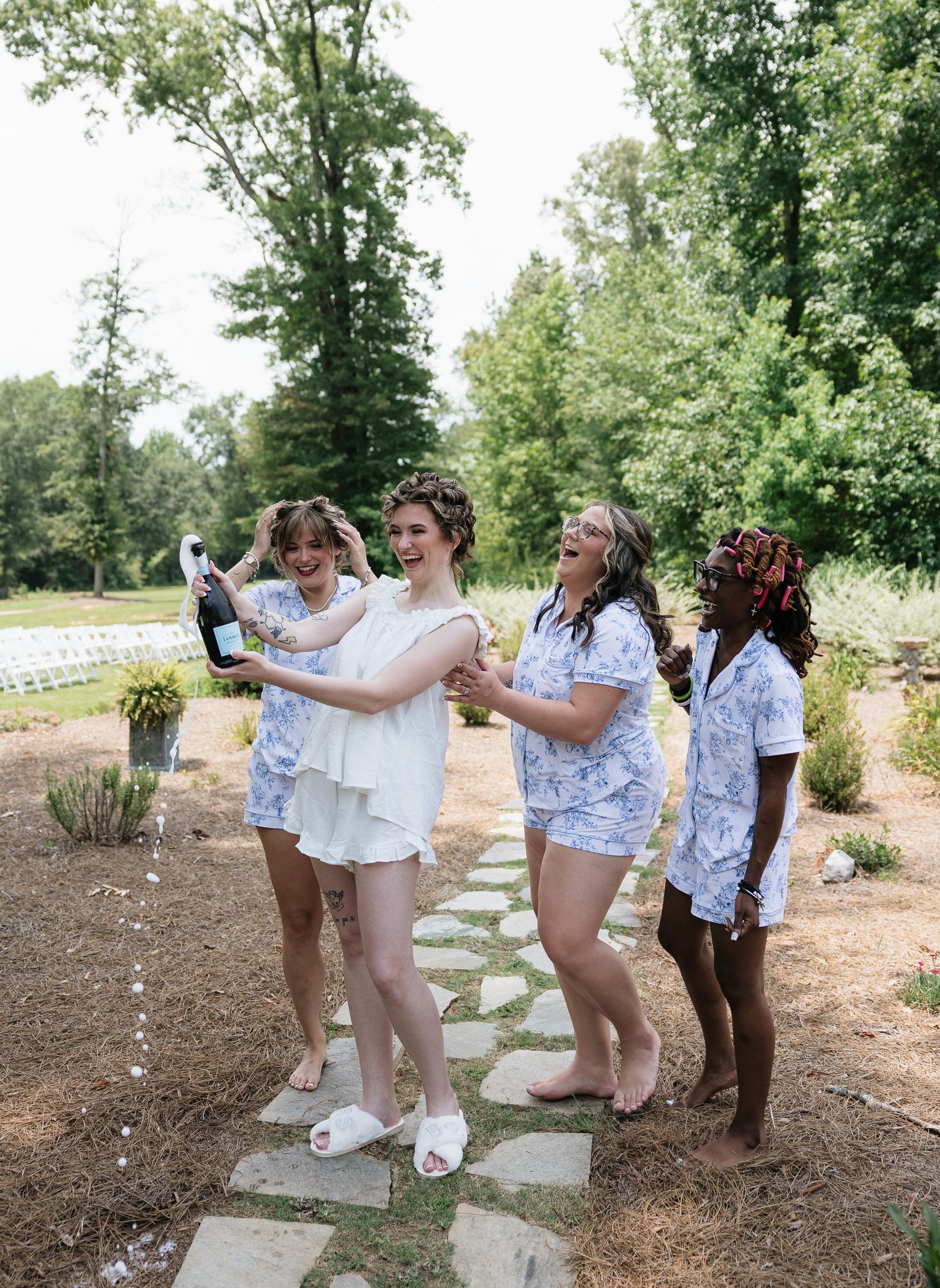 Bride and bridesmaids in matching blue floral pajamas laugh while popping champagne bottle outdoors at wedding venue.