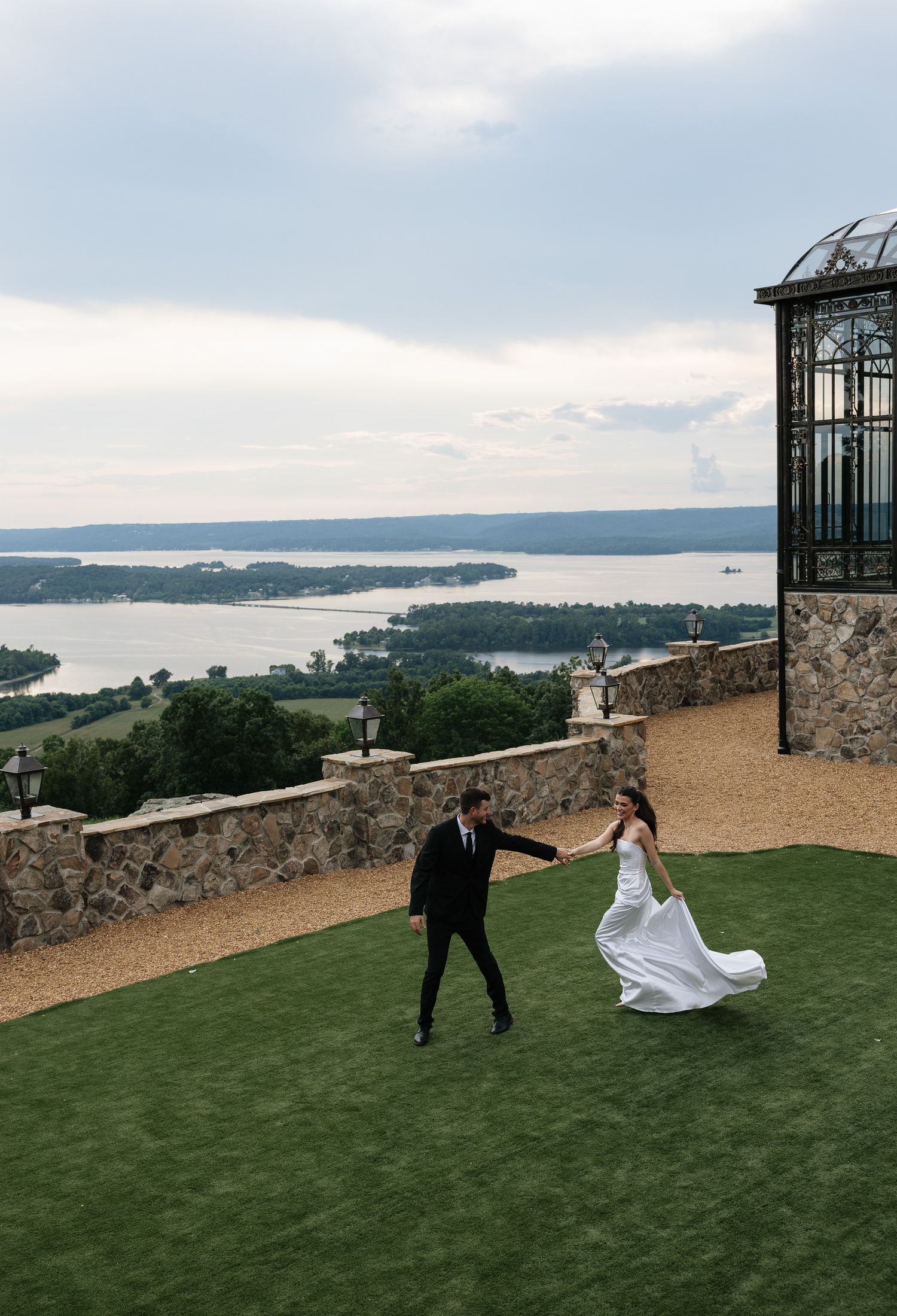 Bride and groom dance on a hilltop terrace overlooking a scenic lake and lush green landscape at an elegant wedding venue.