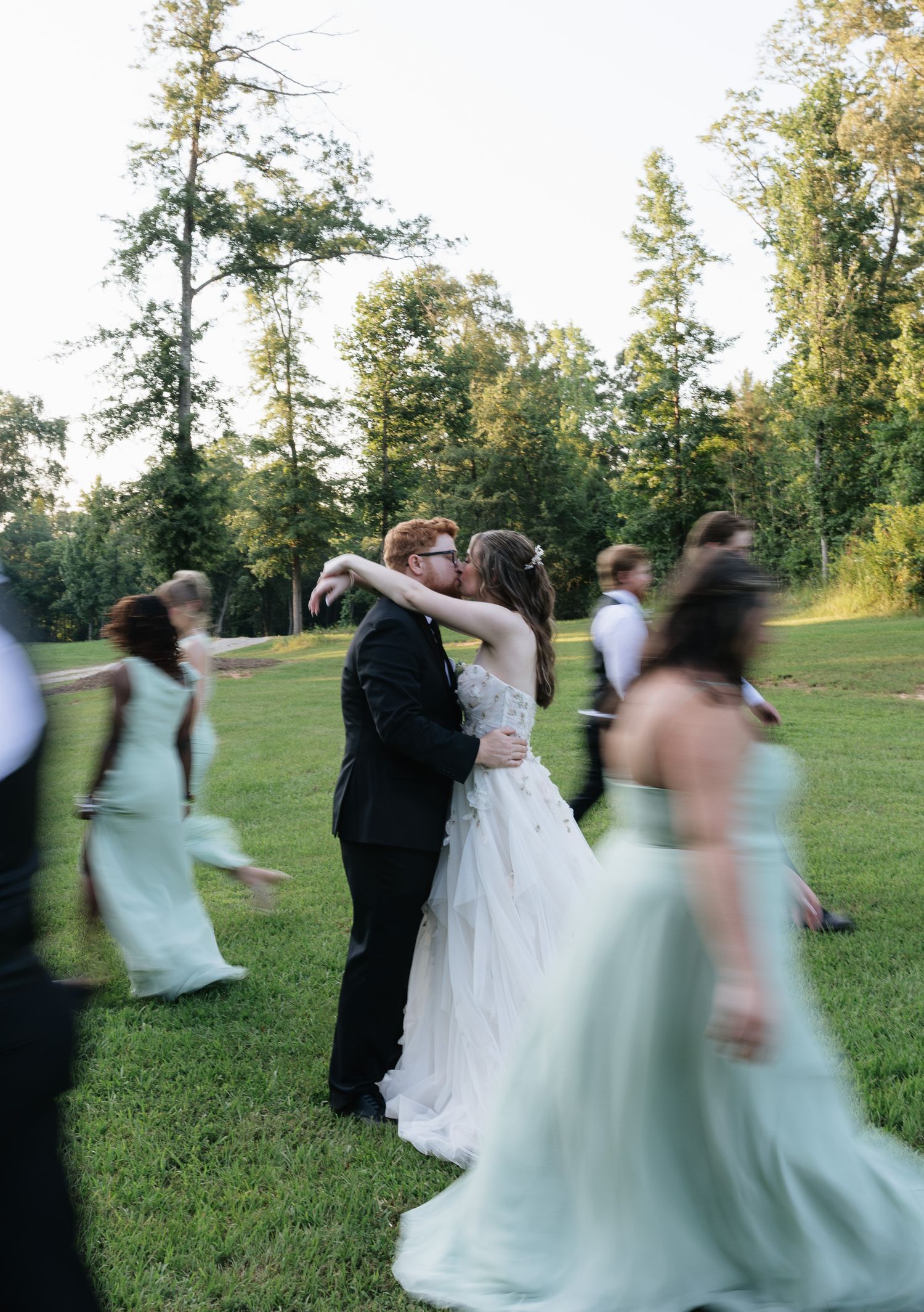 Bride and groom share a kiss surrounded by bridesmaids in mint gowns on a lush green lawn at golden hour.