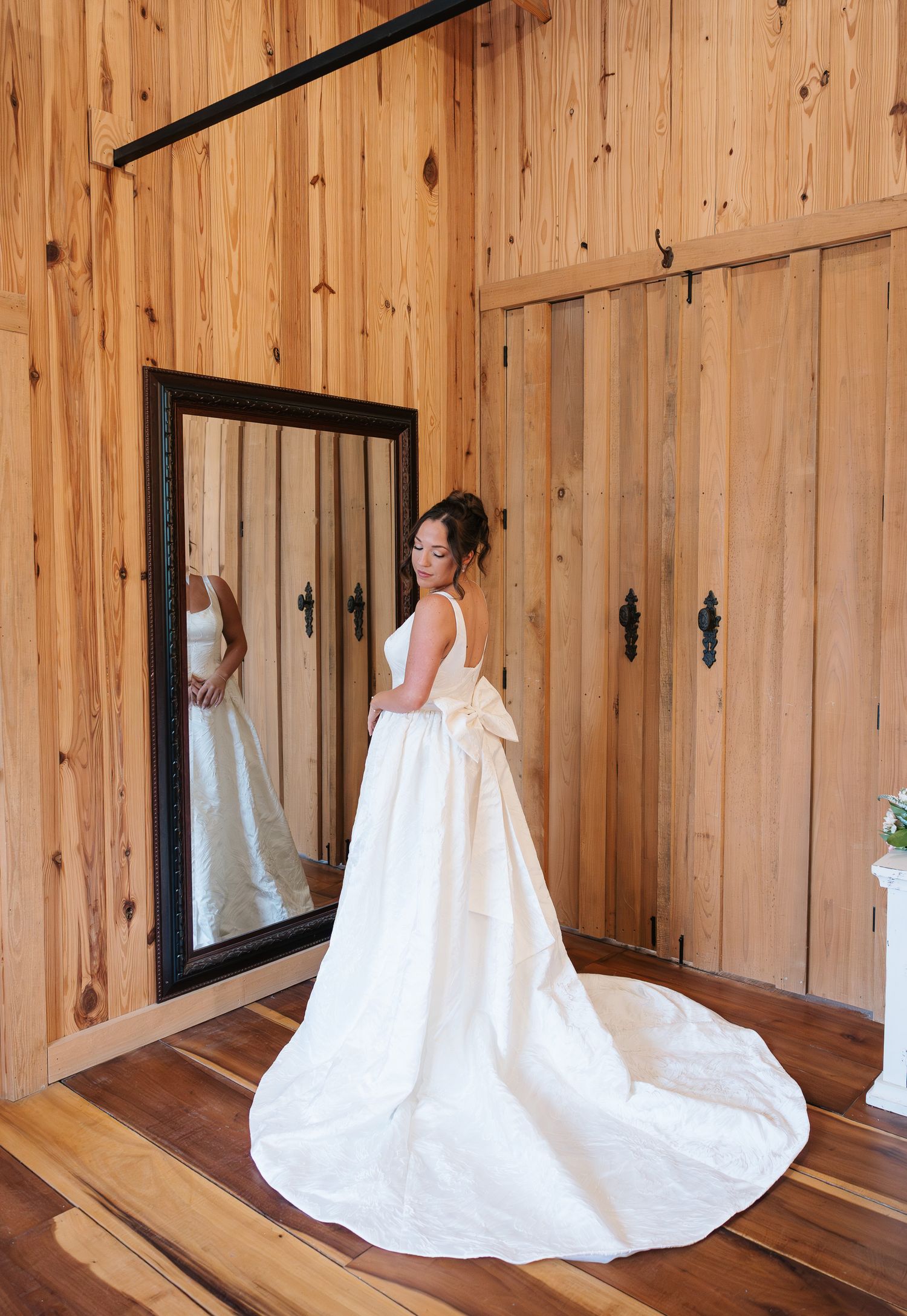 Bride in elegant white wedding gown with bow train stands before rustic wood-paneled barn mirror.