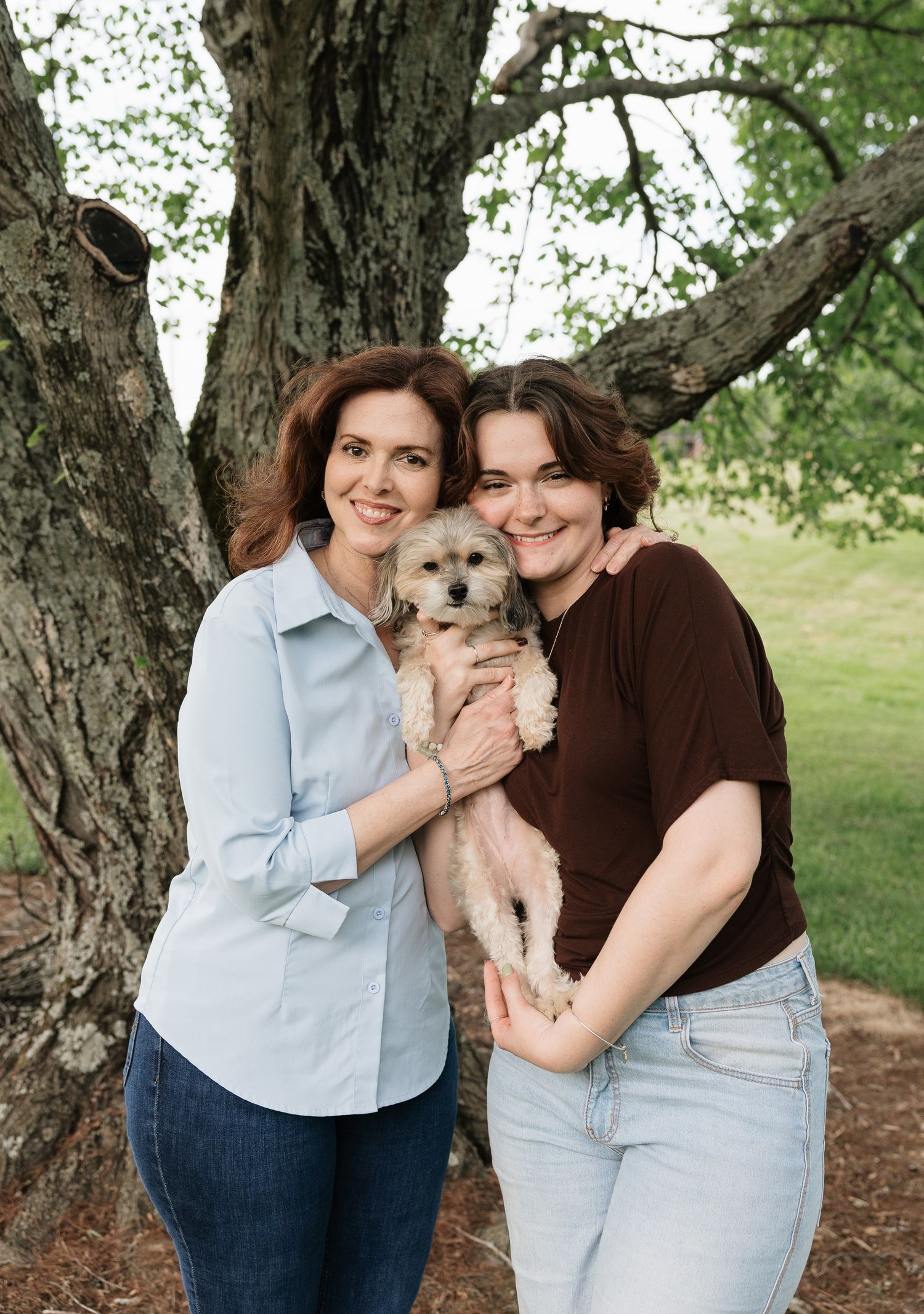 Two smiling women holding a small fluffy dog outdoors near a large tree in a green park setting.