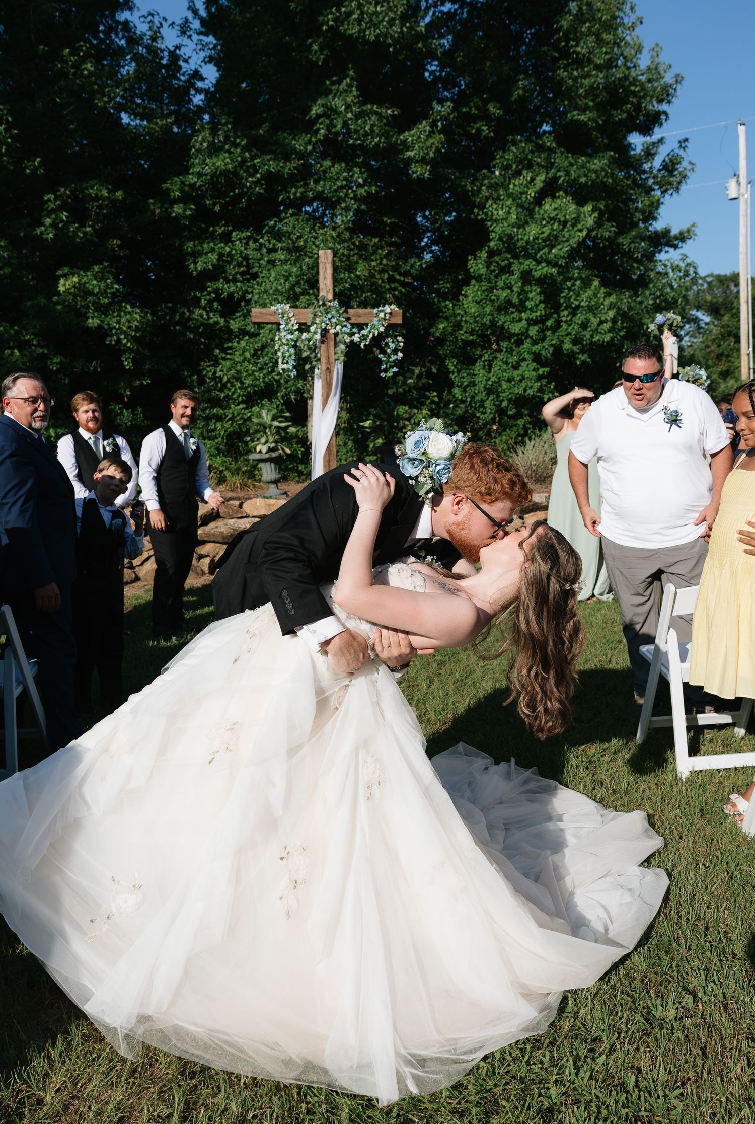 Groom dips bride for a romantic kiss at outdoor wedding ceremony with wooden cross and guests watching.