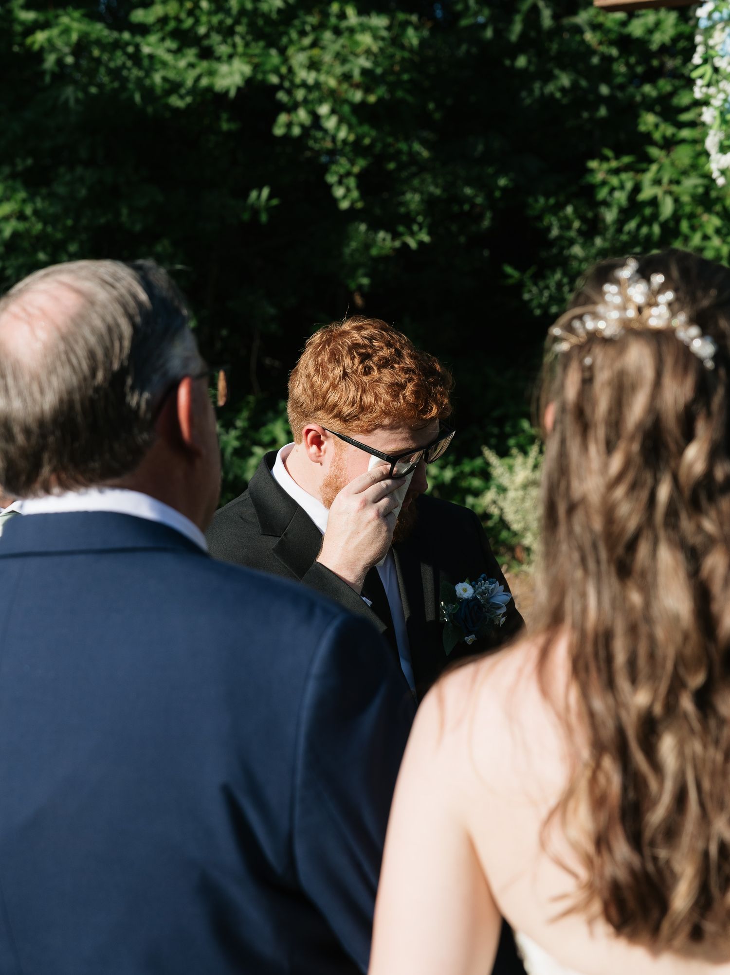 Emotional groom wiping tears during outdoor wedding ceremony with bride and officiant among lush green trees.