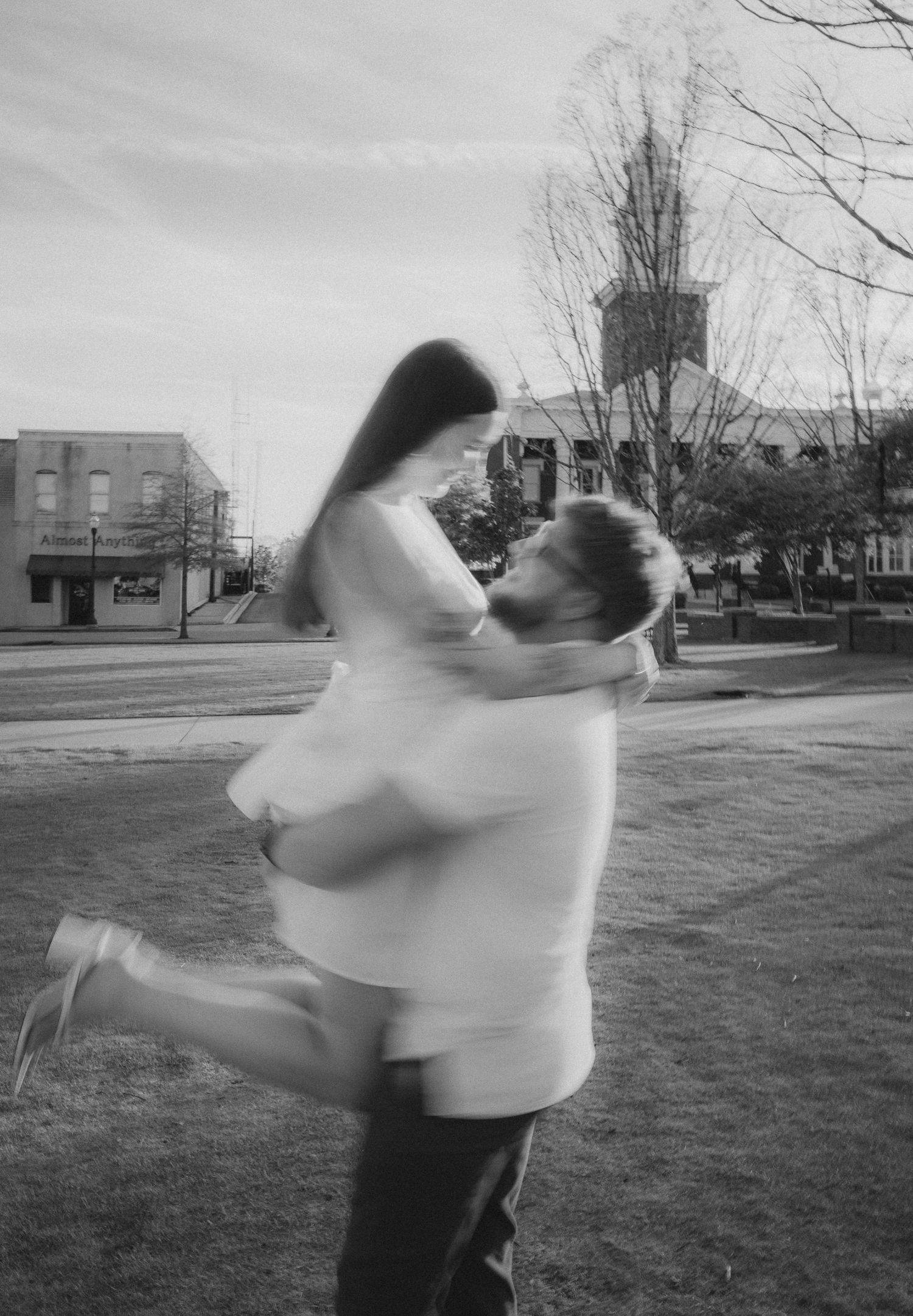 Black and white photo of a couple embracing outdoors, man lifting woman in a romantic spin in a park setting.
