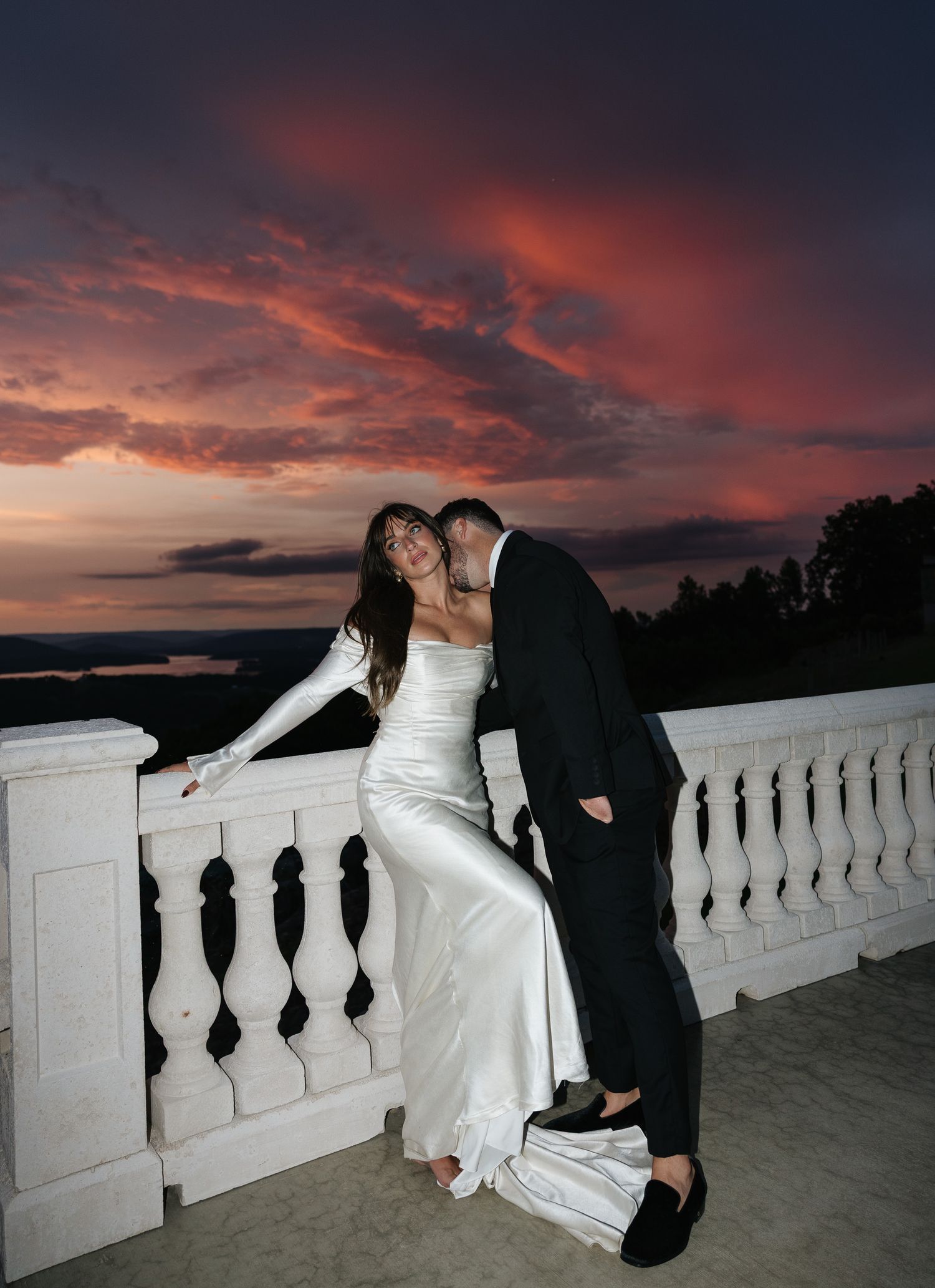 Wedding couple sharing romantic moment on white balustrade terrace under vibrant red sunset sky.
