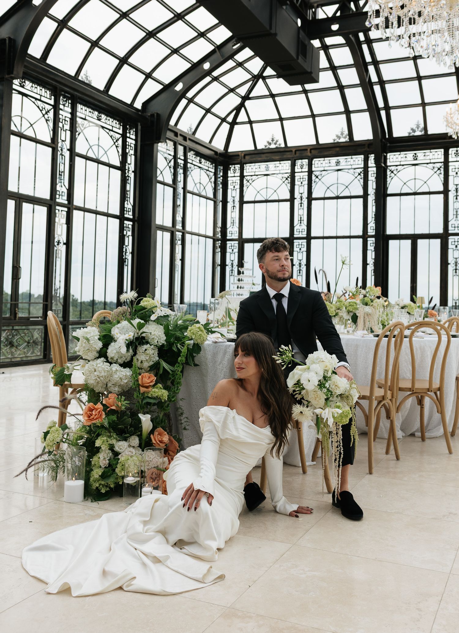 Bride in off-shoulder gown sits on floor with groom in black suit inside elegant glass conservatory with floral arrangements.