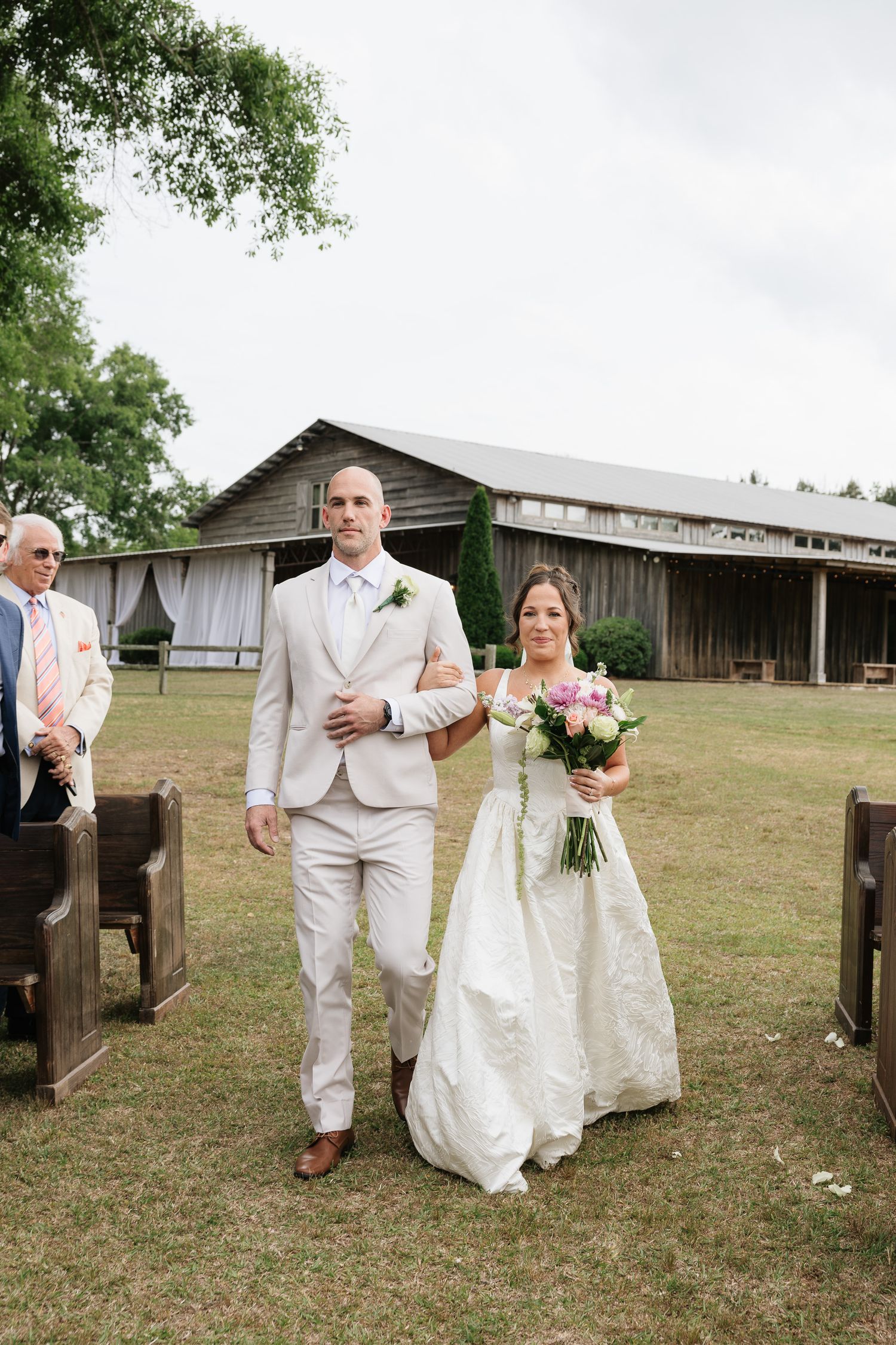 Bride in white gown holding pink bouquet walks down outdoor aisle with groom in beige suit at rustic barn wedding.