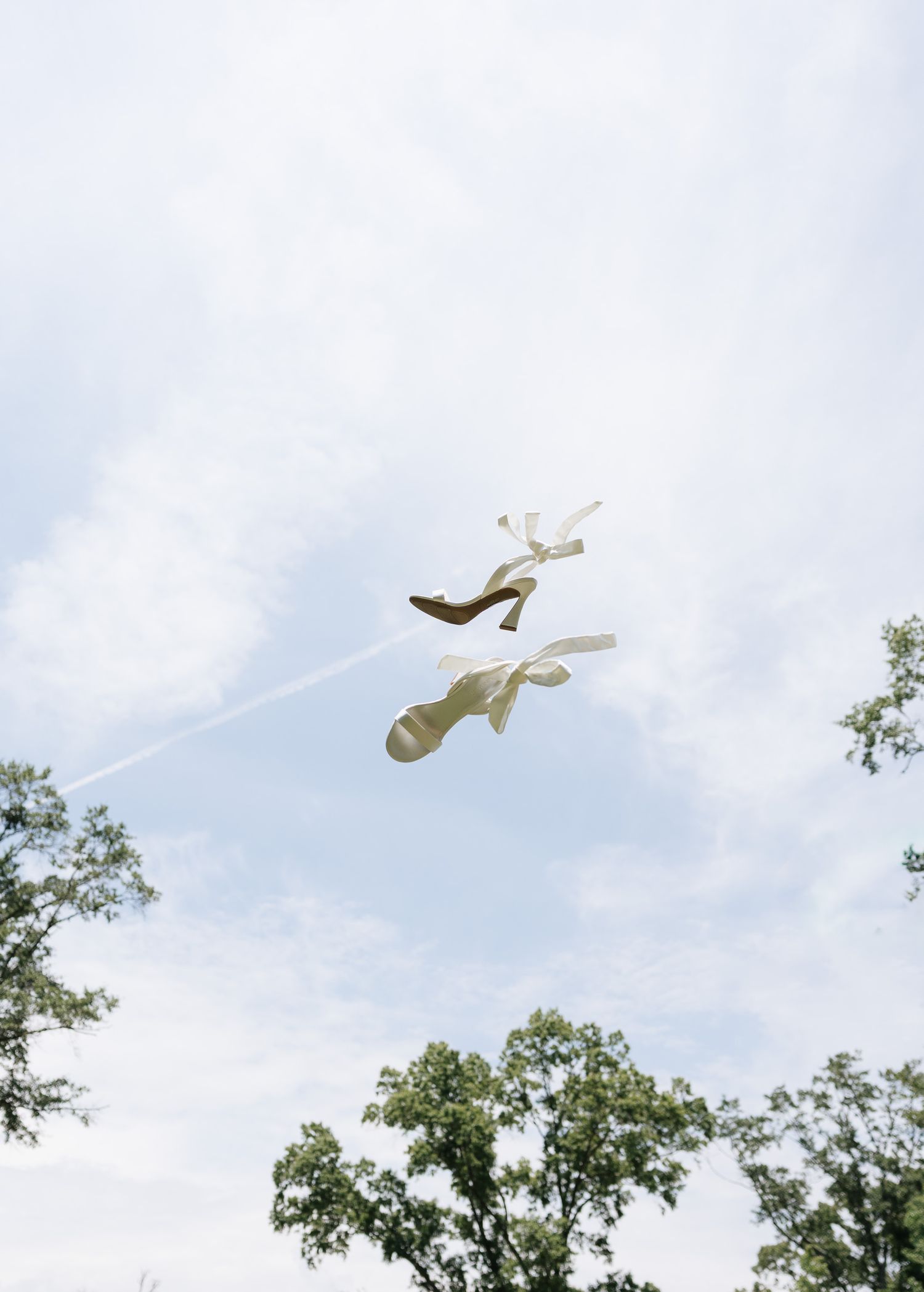Two shoes tossed in the air against a bright cloudy sky with green treetops visible below.