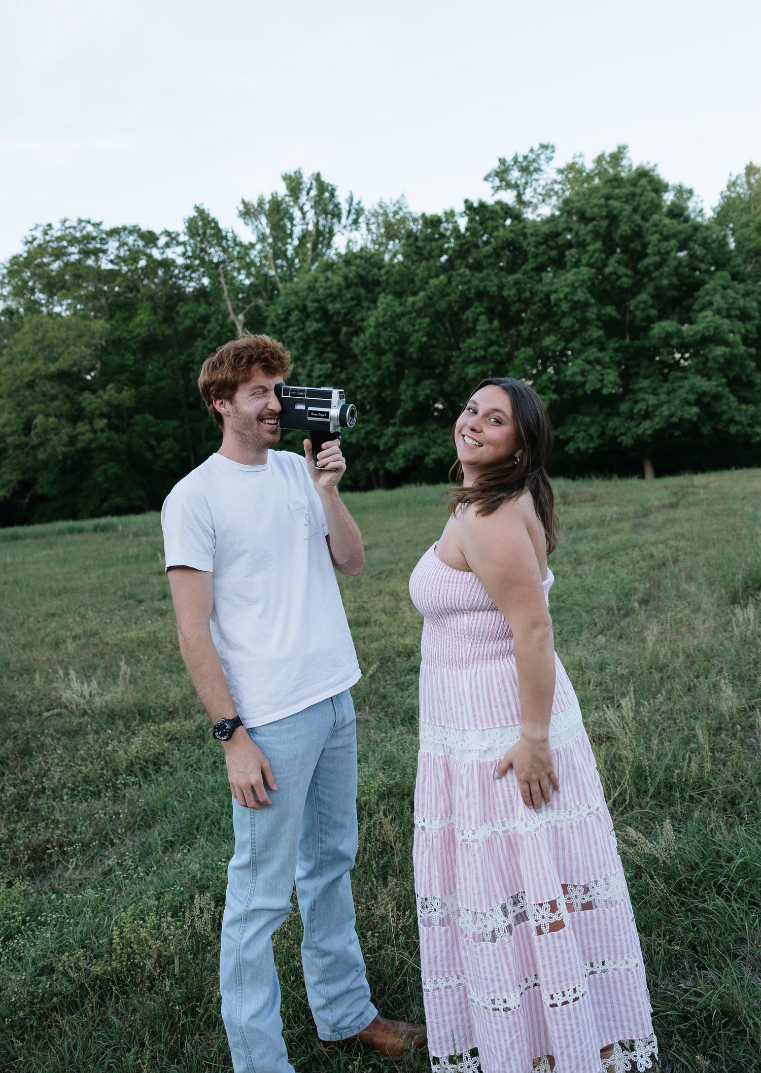 A couple poses in a green field, man in white tee holding vintage camera, woman in pink lace maxi dress smiling.