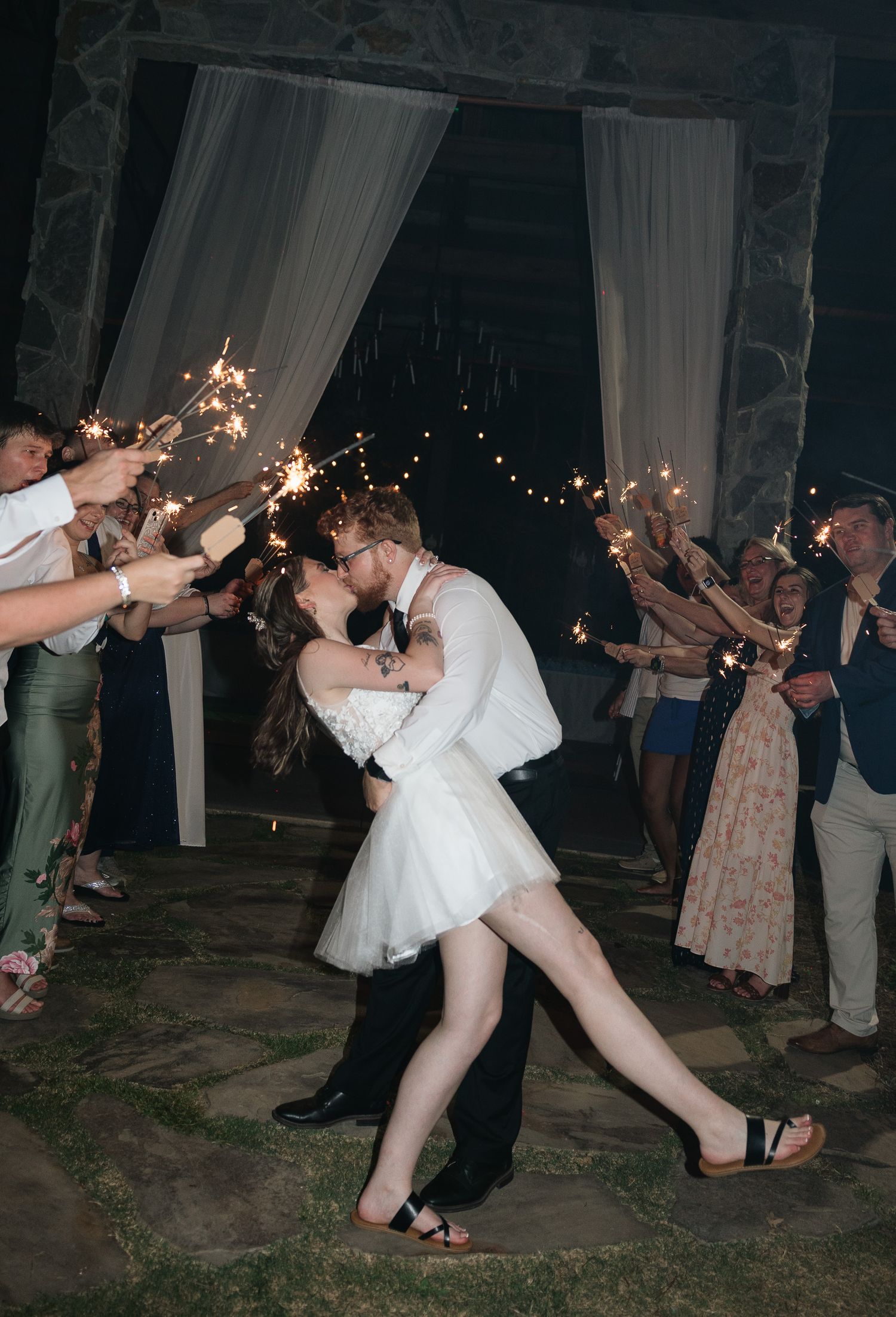 Bride and groom share romantic kiss during sparkler send-off exit at night, bride in short white dress lifted by groom.