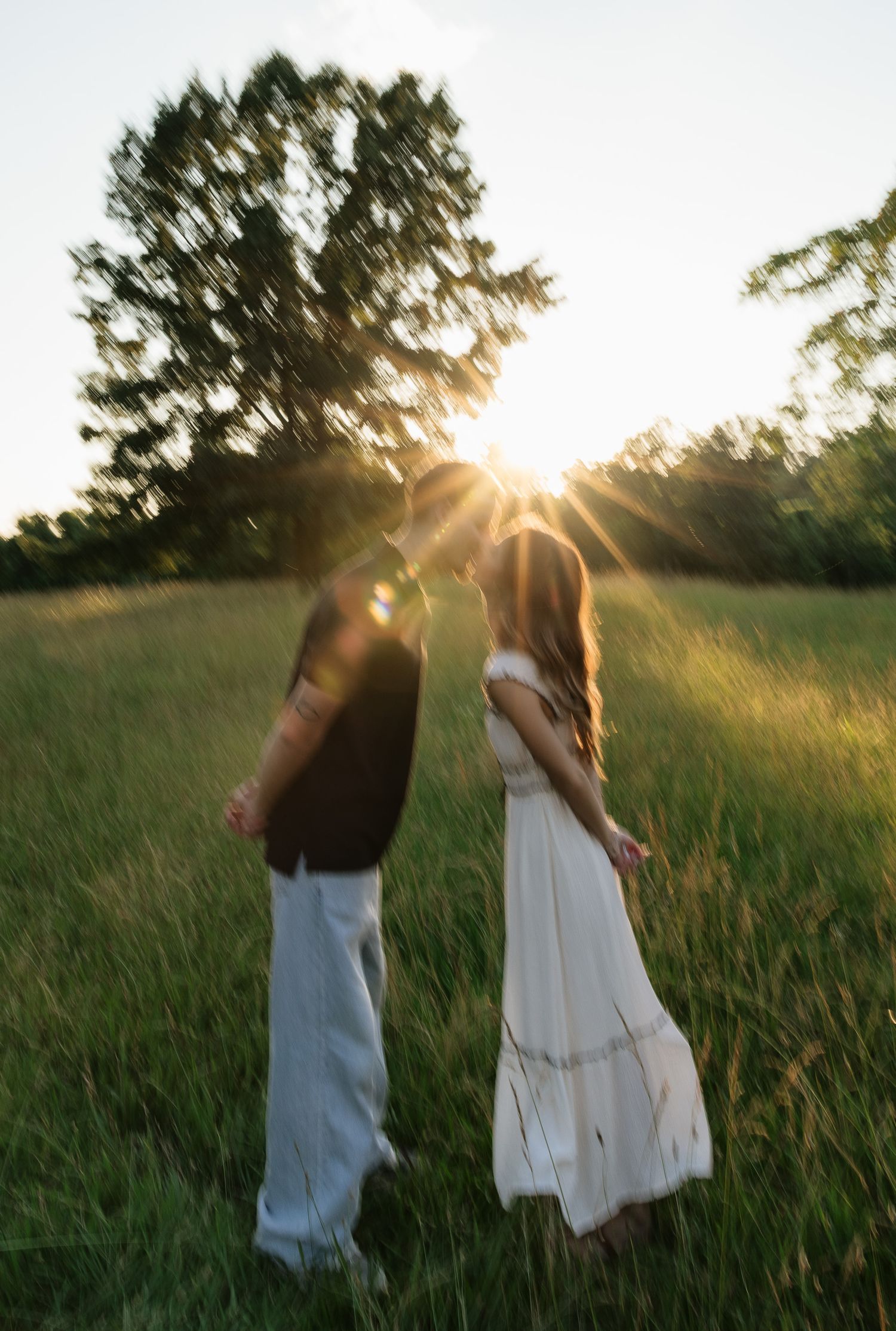 Couple standing in a sunlit meadow at golden hour, backlit by sun rays through trees, woman in white dress.