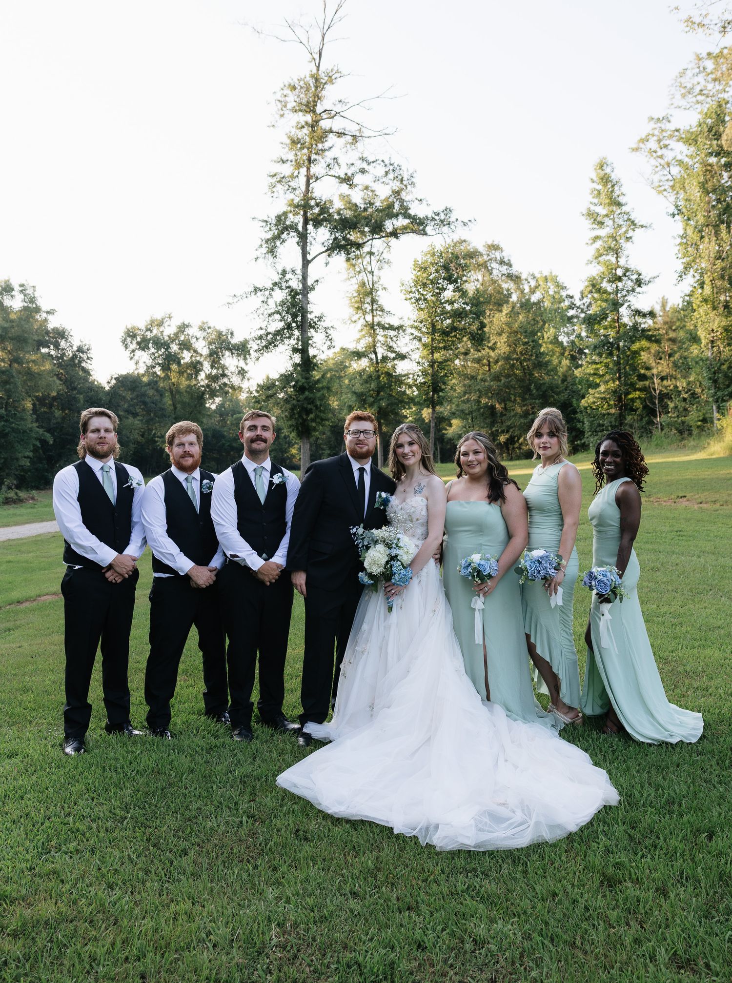 Wedding party poses outdoors on green grass, groomsmen in black suits and bridesmaids in sage green dresses with bride and groom centered.