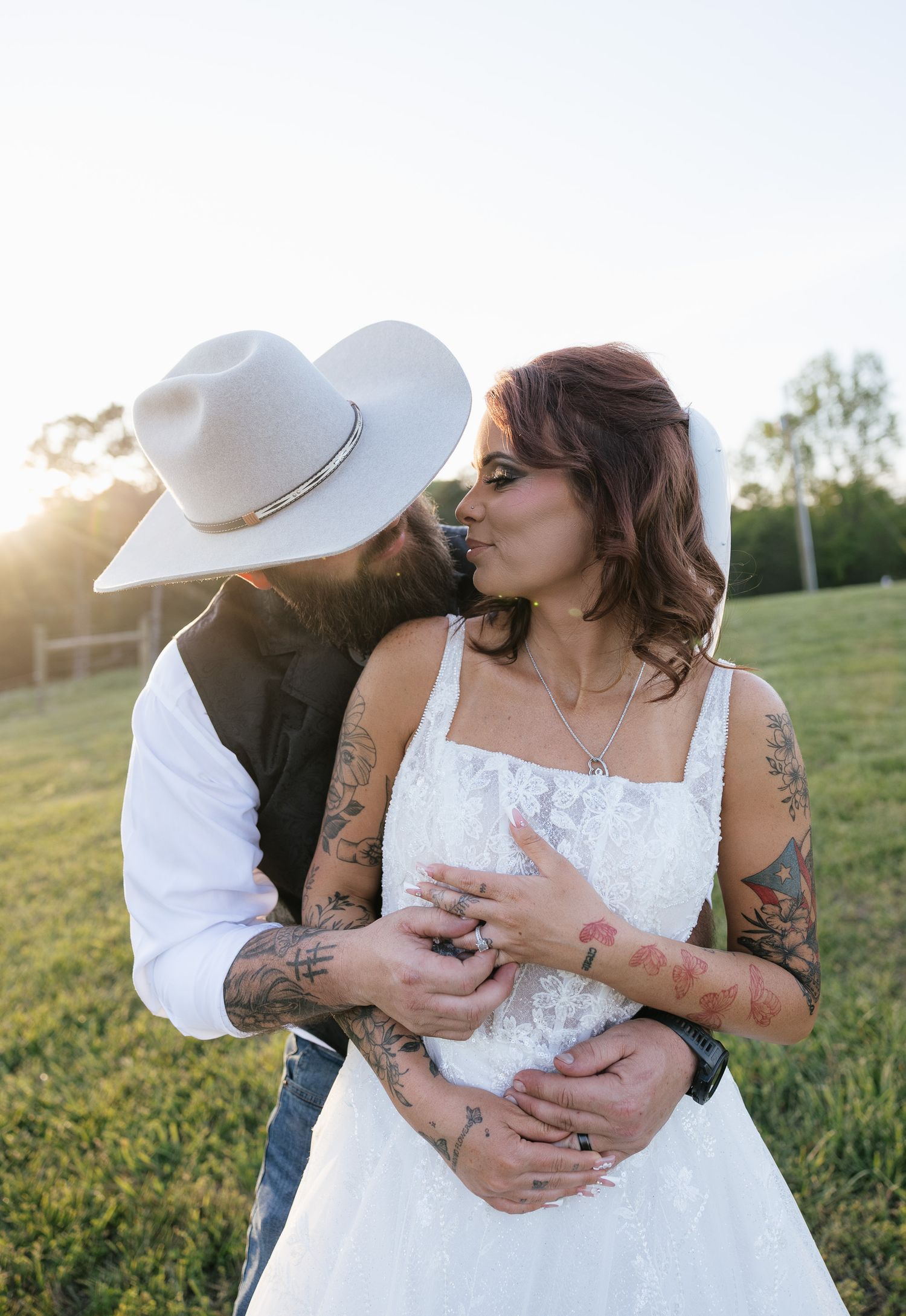 Tattooed couple sharing a romantic moment at golden hour, bride in white lace gown and groom in white cowboy hat outdoors.