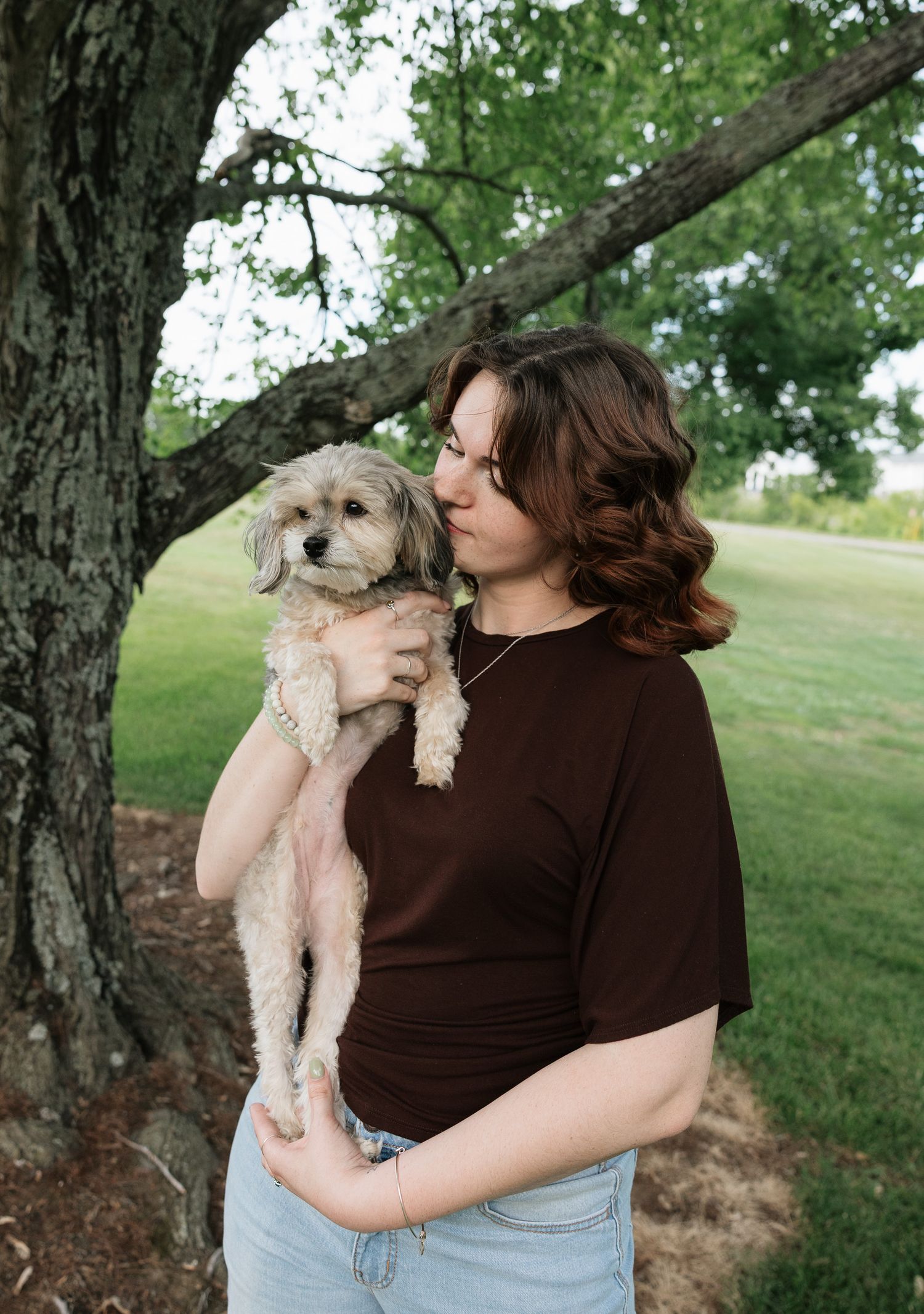 Young woman in brown top holding a fluffy small dog outdoors near a large tree in a green park.