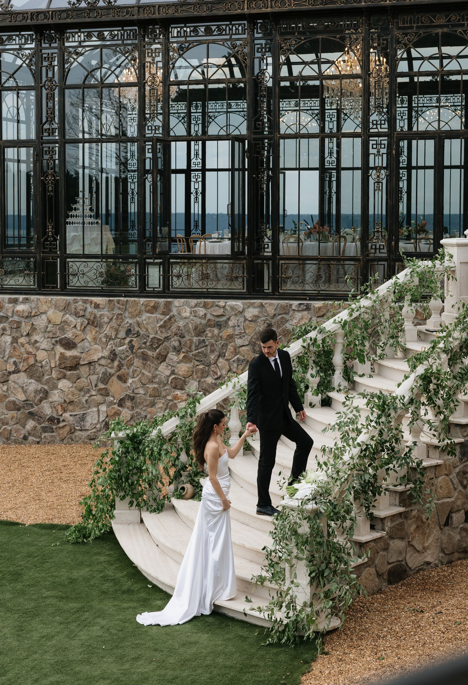 Bride and groom on vine-covered stone stairs outside elegant glass conservatory venue.