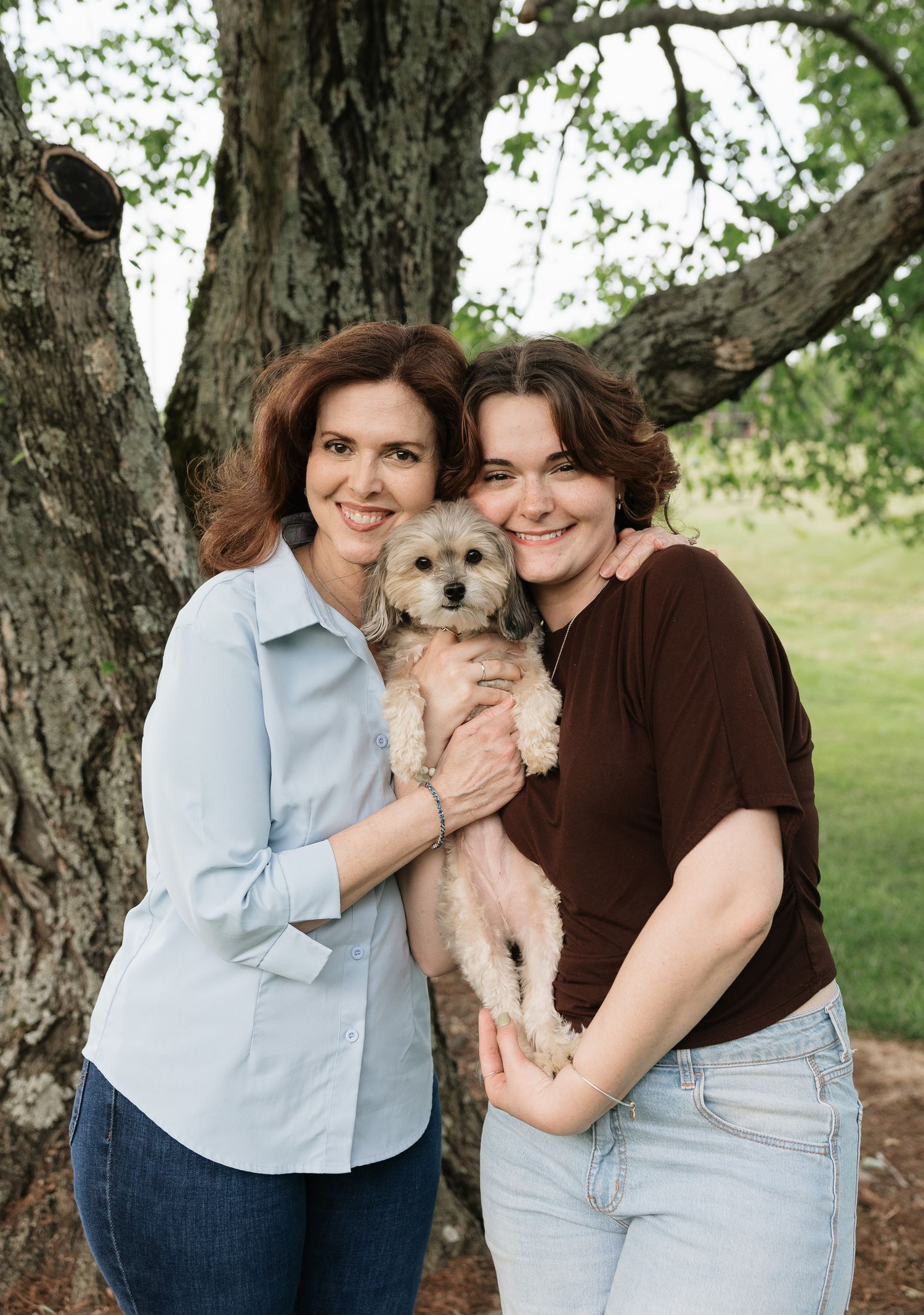 Two women smiling outdoors holding a small fluffy dog near a large tree.