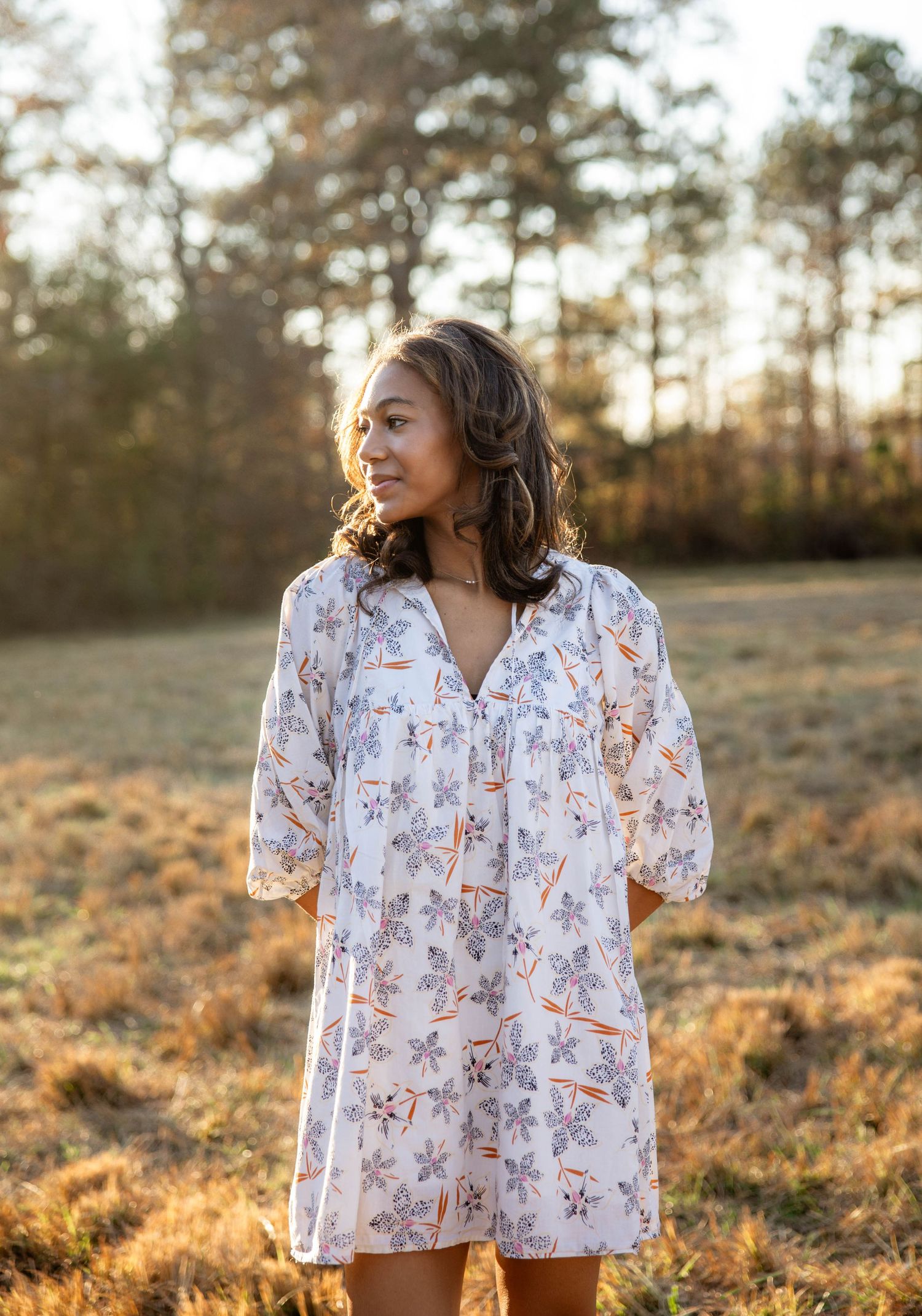 A person in a white floral dress poses outdoors during golden hour sunset in a grassy field.