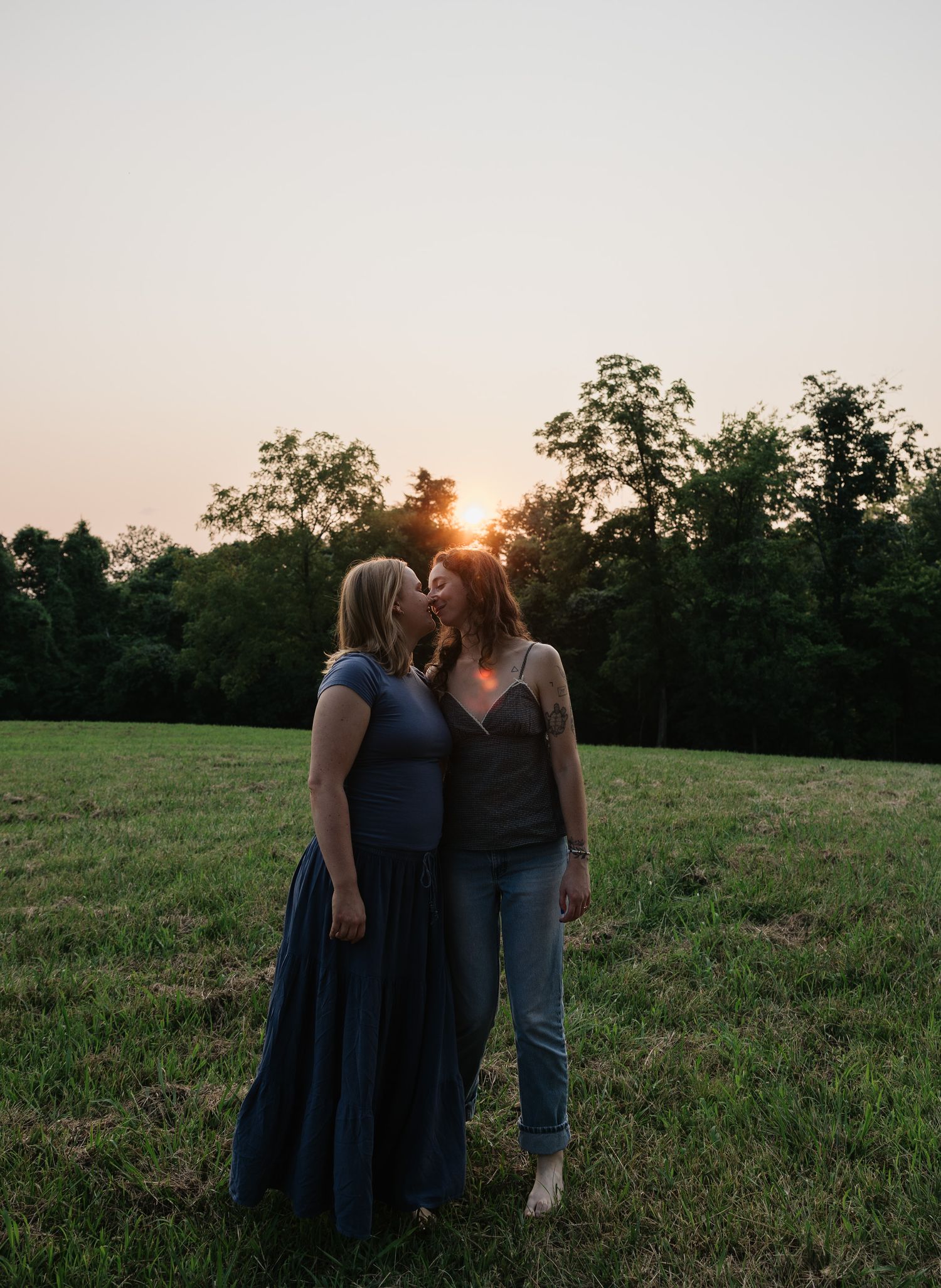 Two women sharing a tender kiss at golden hour sunset in a lush green field, one in a blue gown and one in jeans.