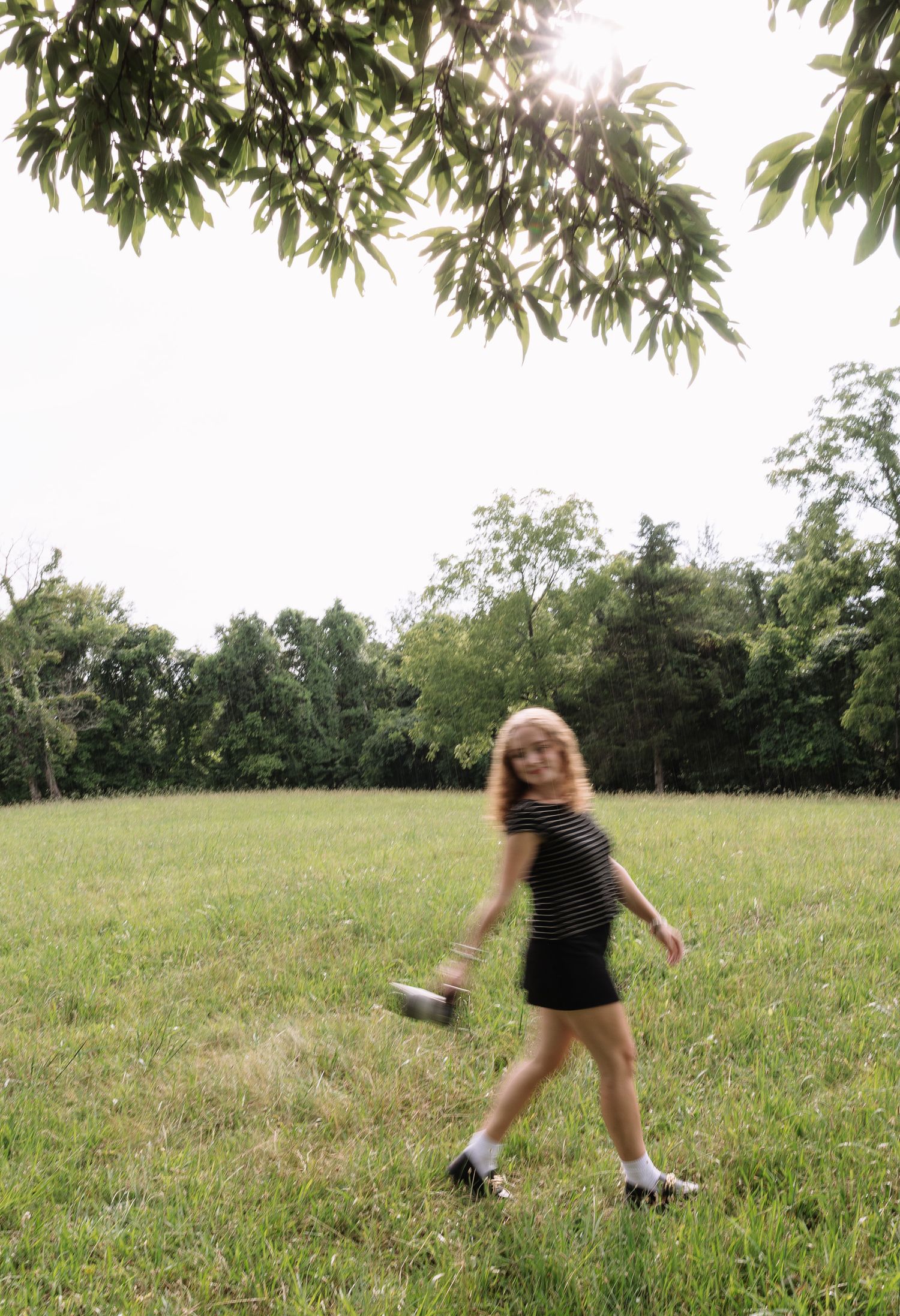 Young blonde woman in striped dress walking across green field, holding a can, with trees in background.