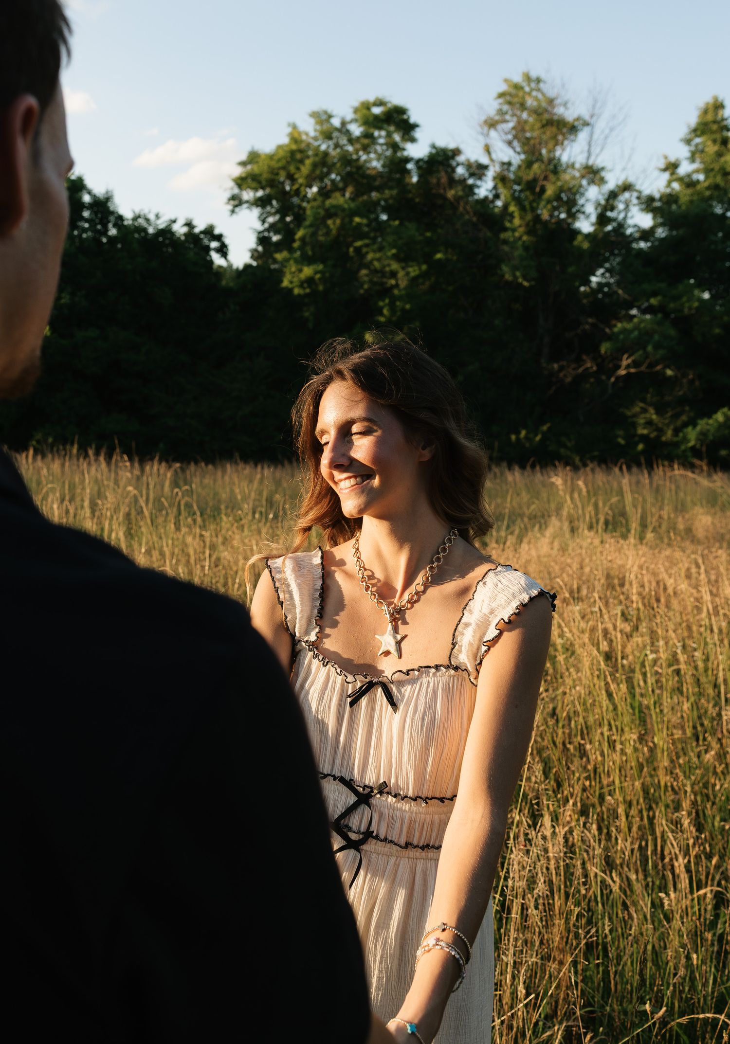 A smiling woman in a white dress with black trim stands in a golden field at sunset.