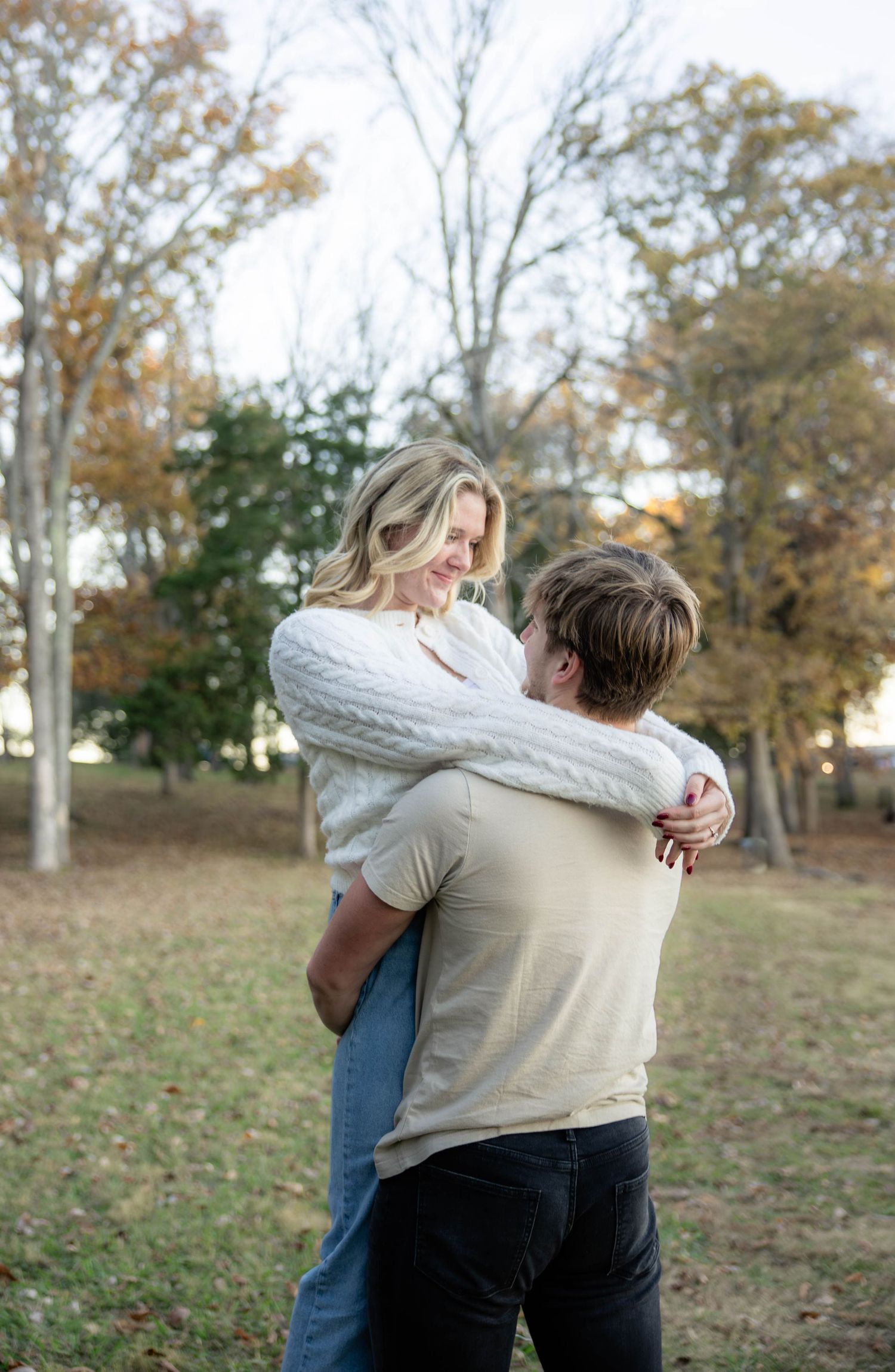 Two people embrace while standing in a park with autumn trees in the background.