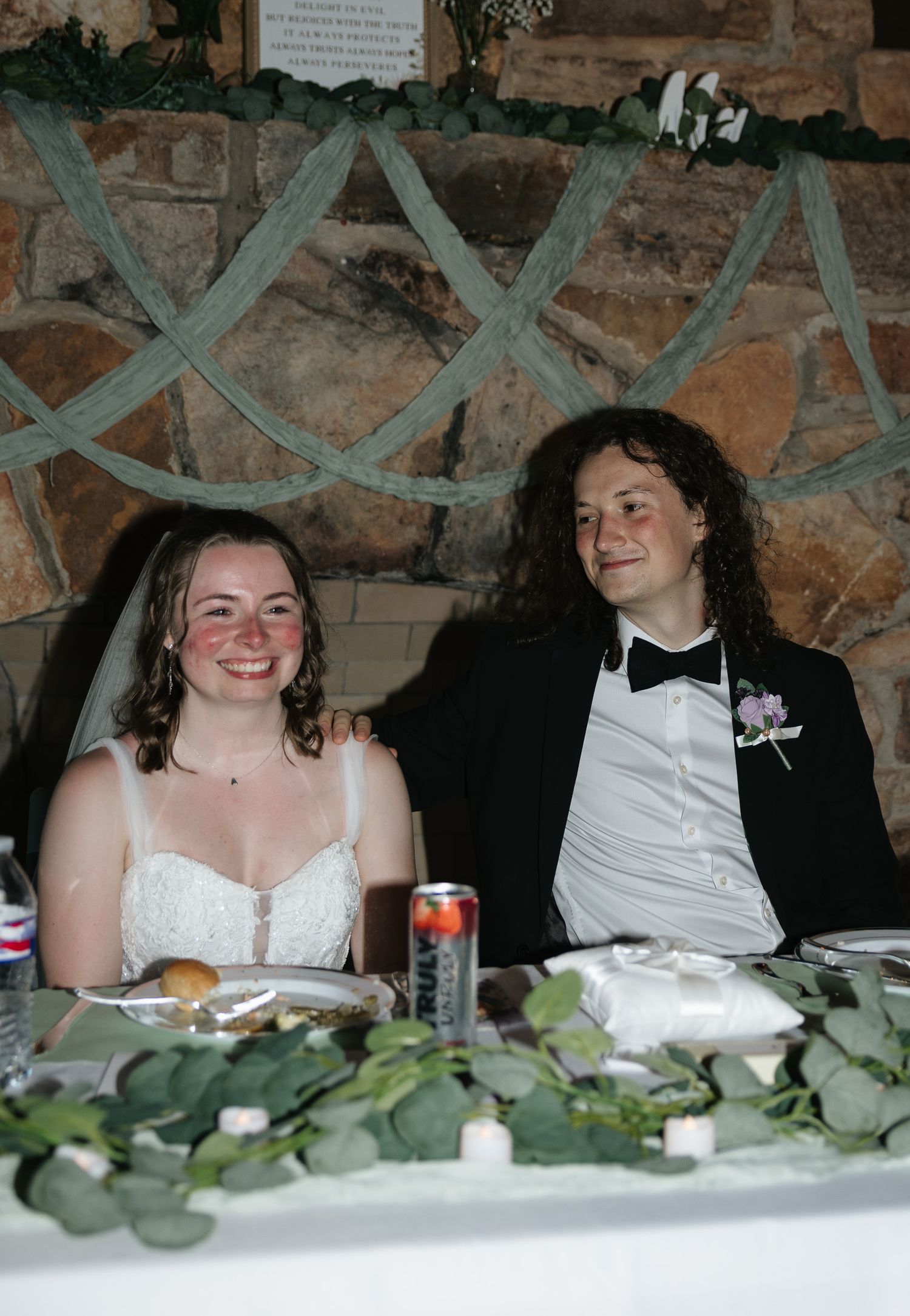 Bride and groom sitting together at wedding reception table decorated with greenery, stone fireplace backdrop.
