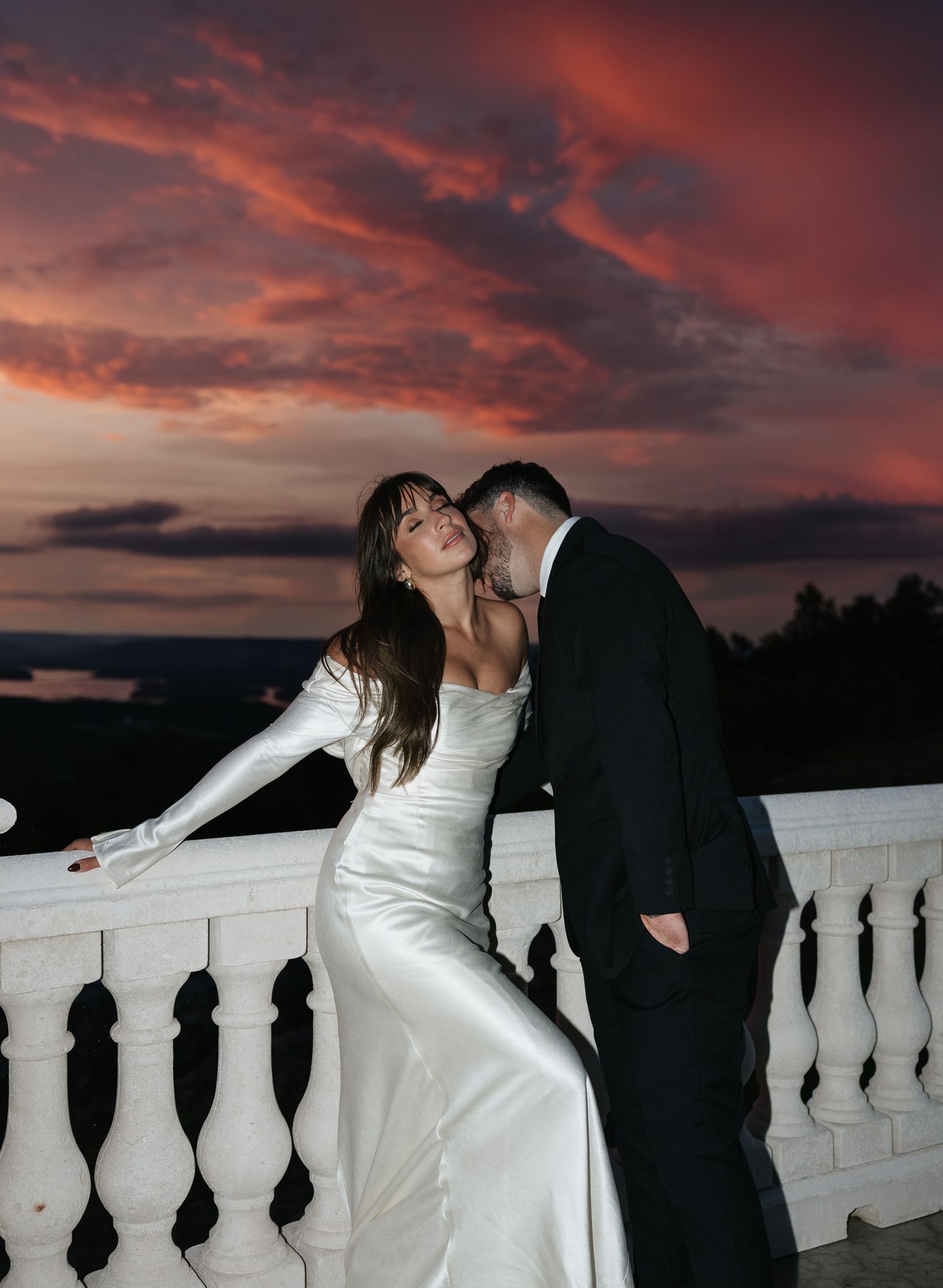 Bride in white satin gown and groom embracing on terrace balcony beneath dramatic crimson sunset.