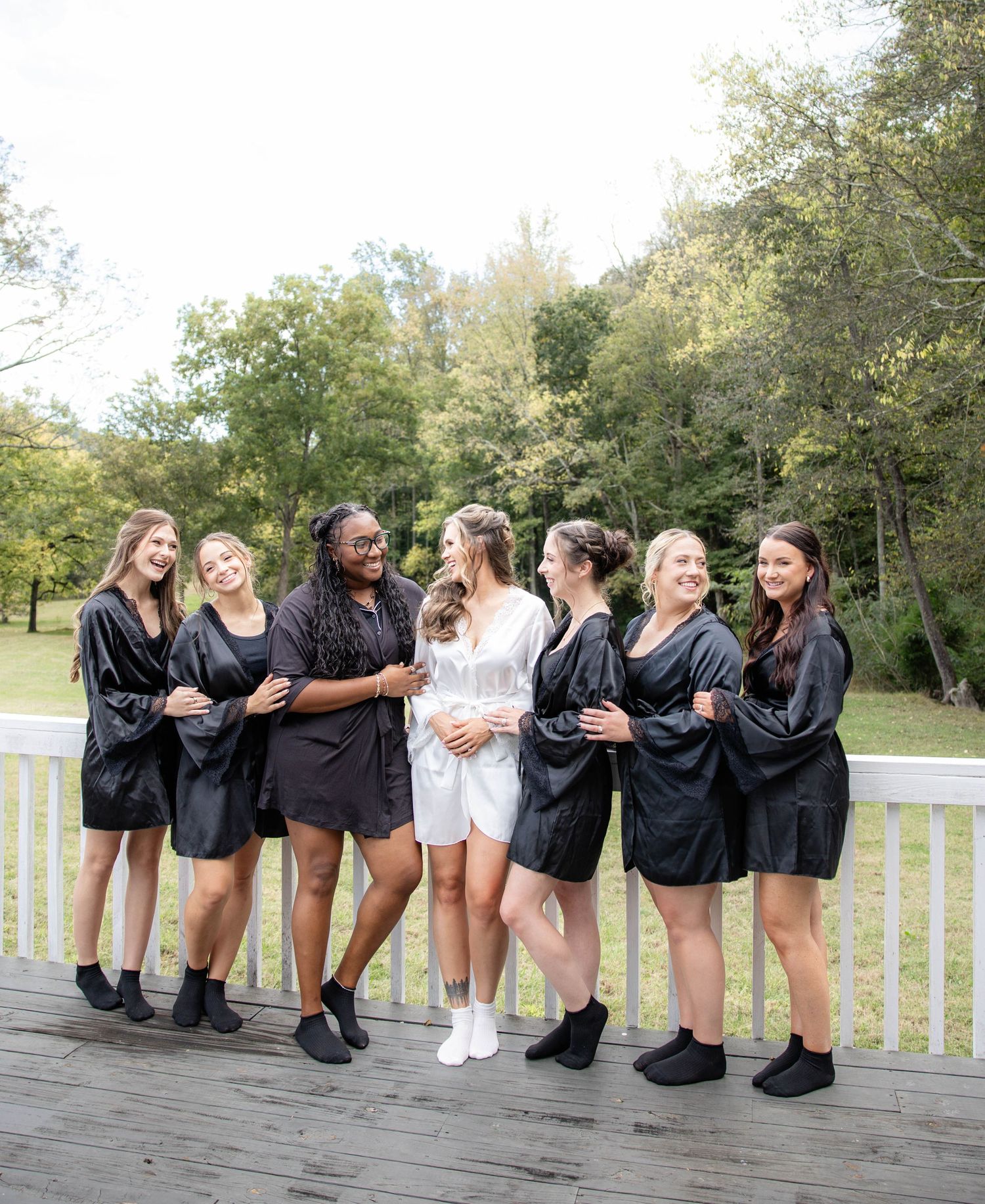 Bridesmaids in black robes pose playfully on a wooden deck showing off their matching outfits.