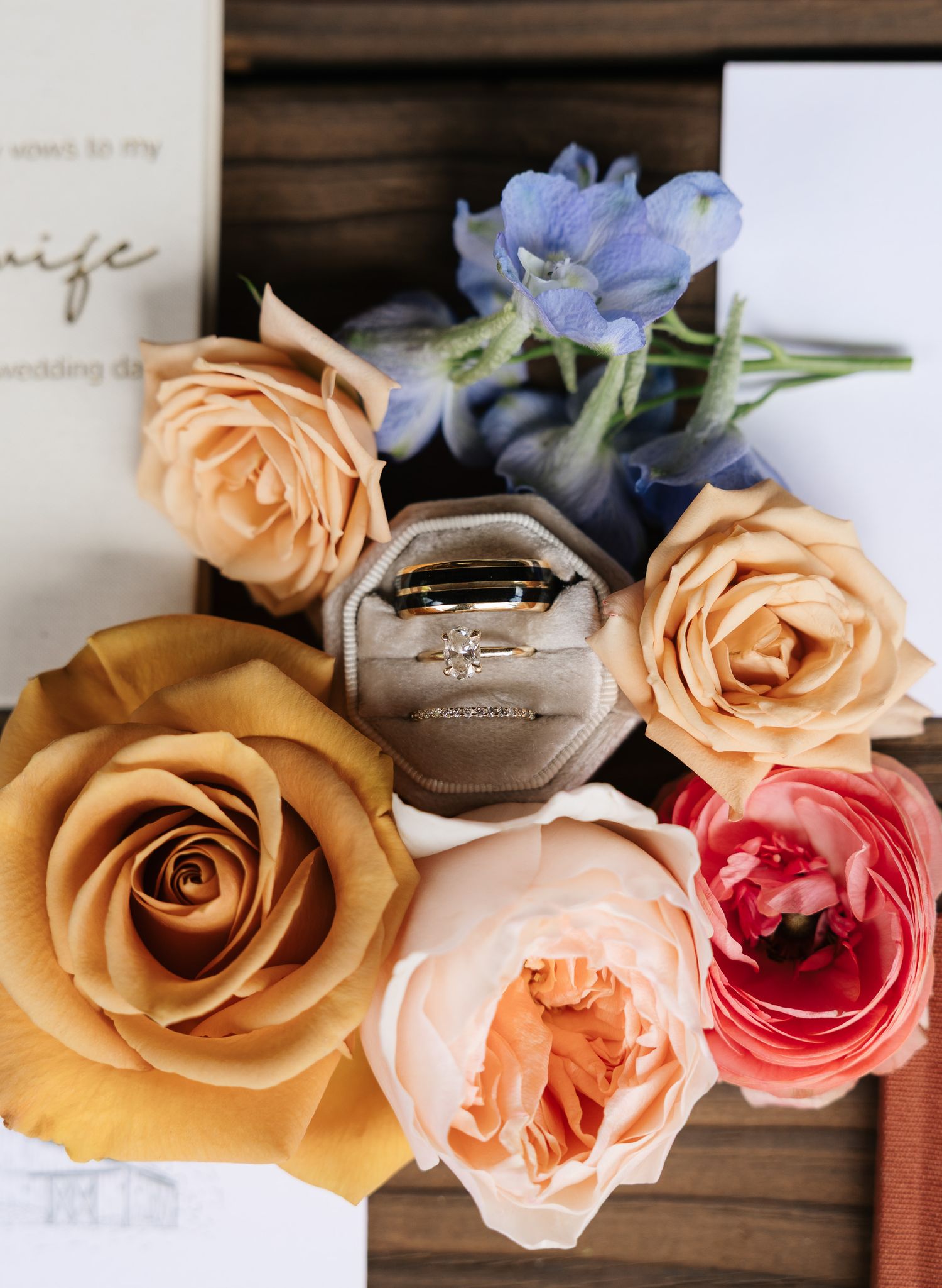 Wedding rings in a velvet box surrounded by peach, coral, and blue flowers on a rustic wooden tray.