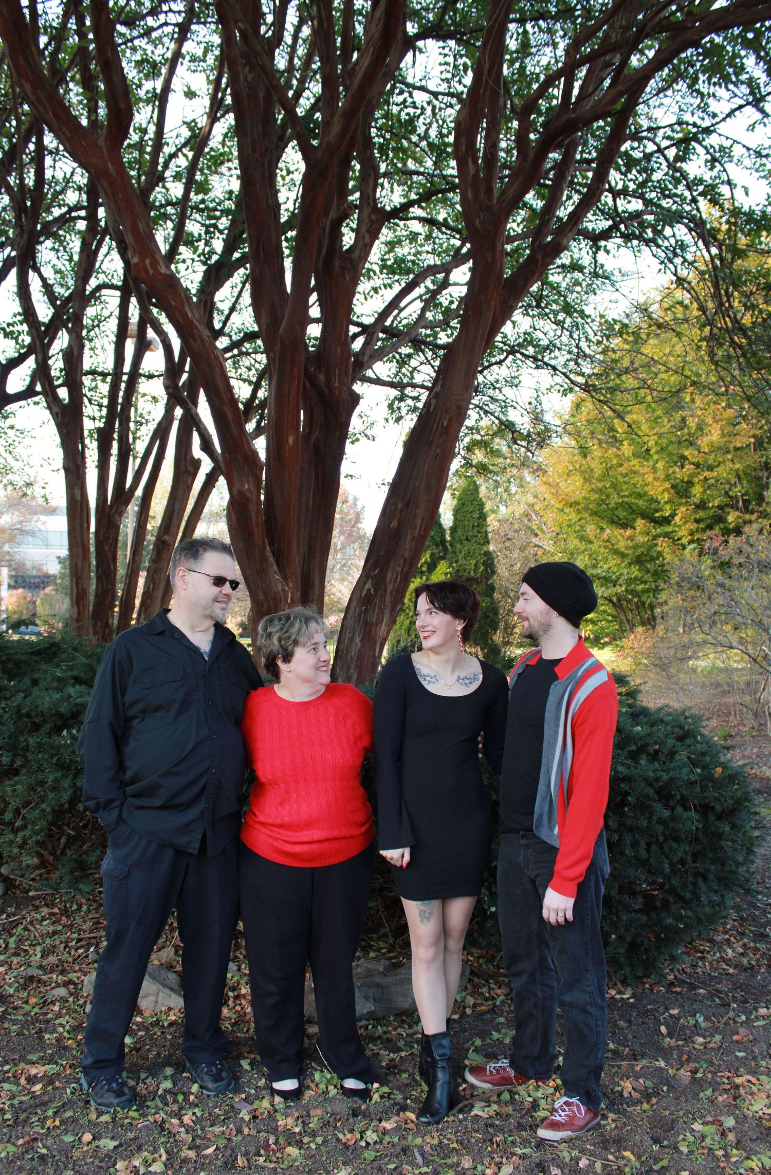 A group in black outfits and one red top pose playfully against trees in an autumn setting.