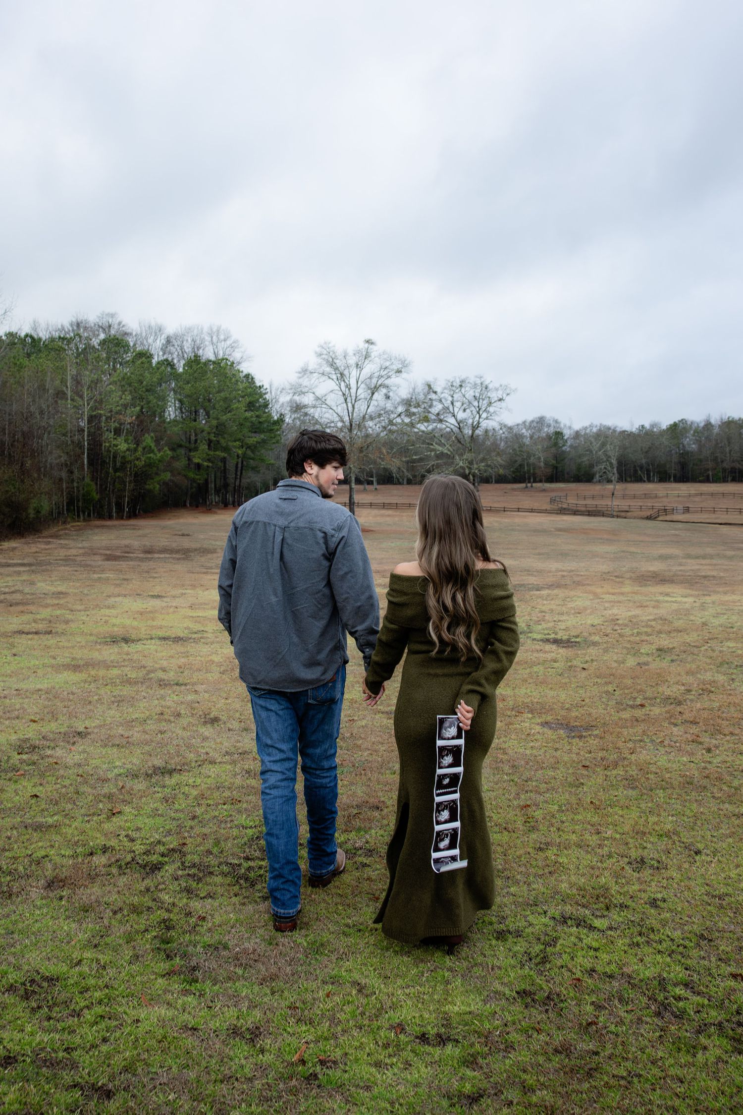 Couple holding hands in a field, woman in olive green dress displaying ultrasound photos, pregnancy announcement outdoors.