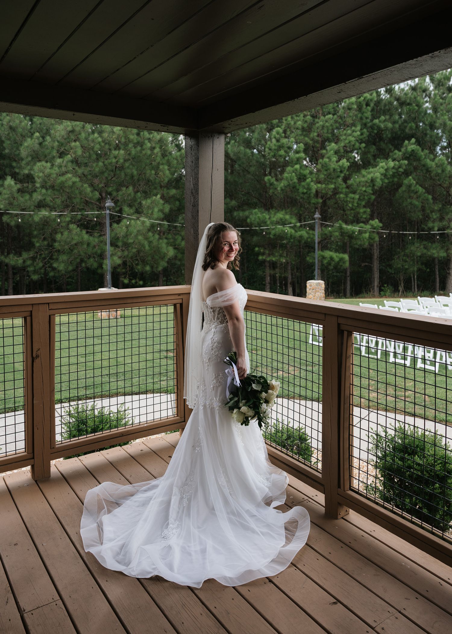 Elegant bride in fitted white gown with long train and veil holding bouquet on wooden deck overlooking outdoor venue.