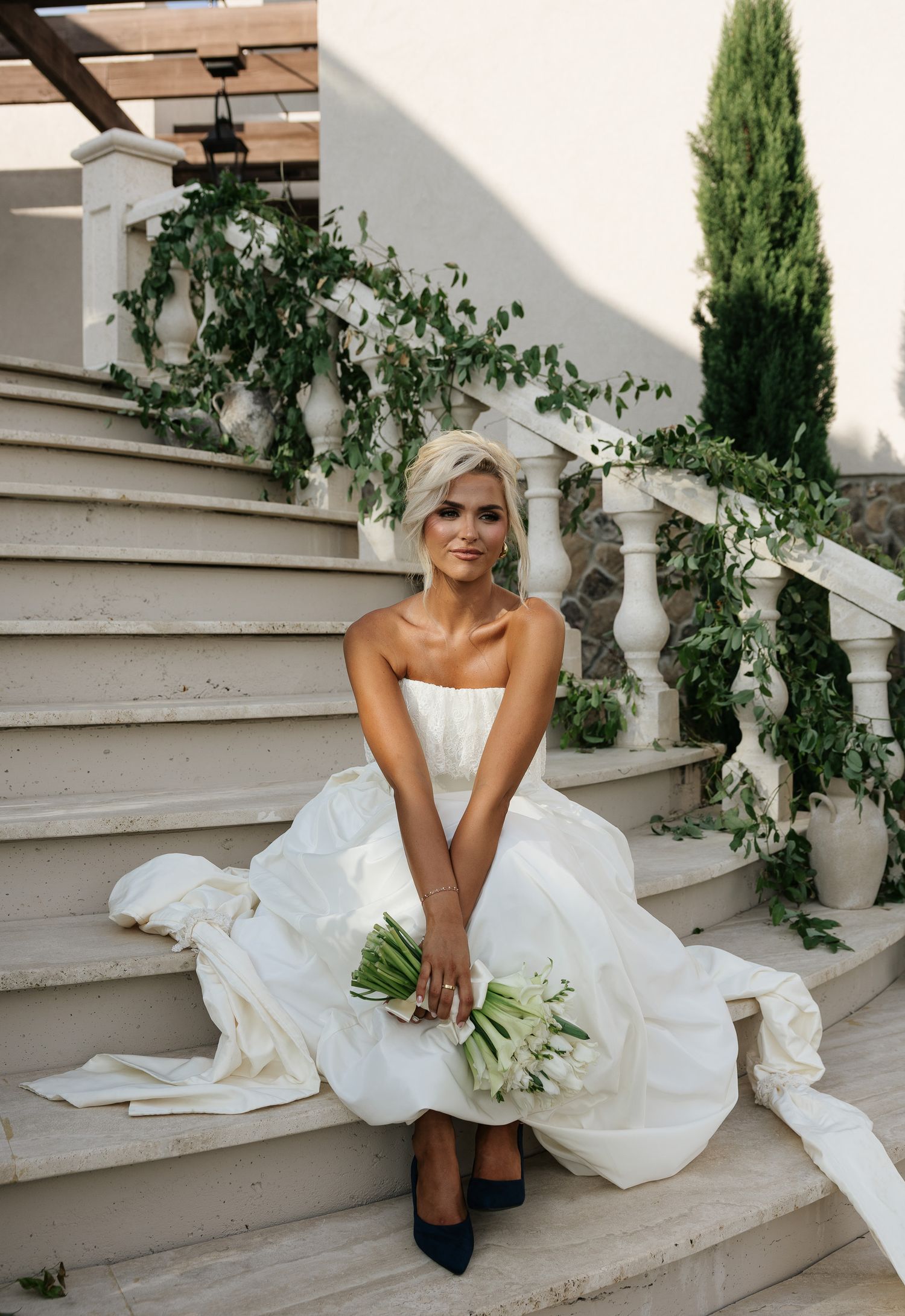Bride in strapless white ballgown sits on stone steps holding white bouquet with navy heels at outdoor venue.