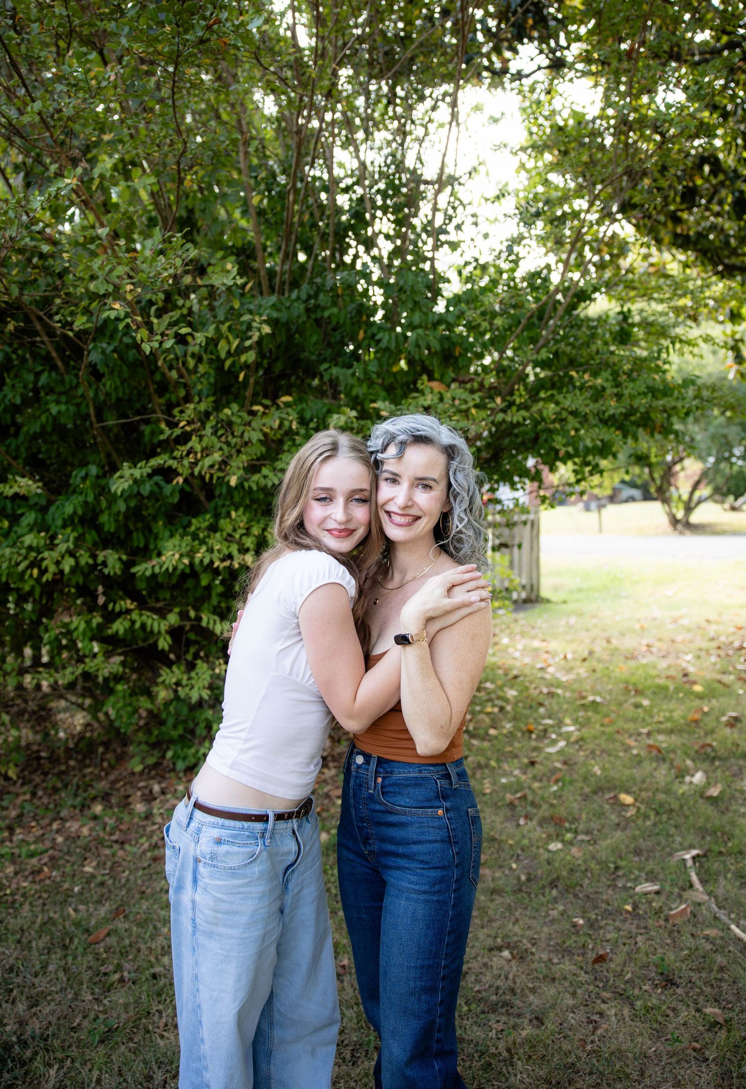 Two friends share a warm embrace outdoors against a leafy green background.