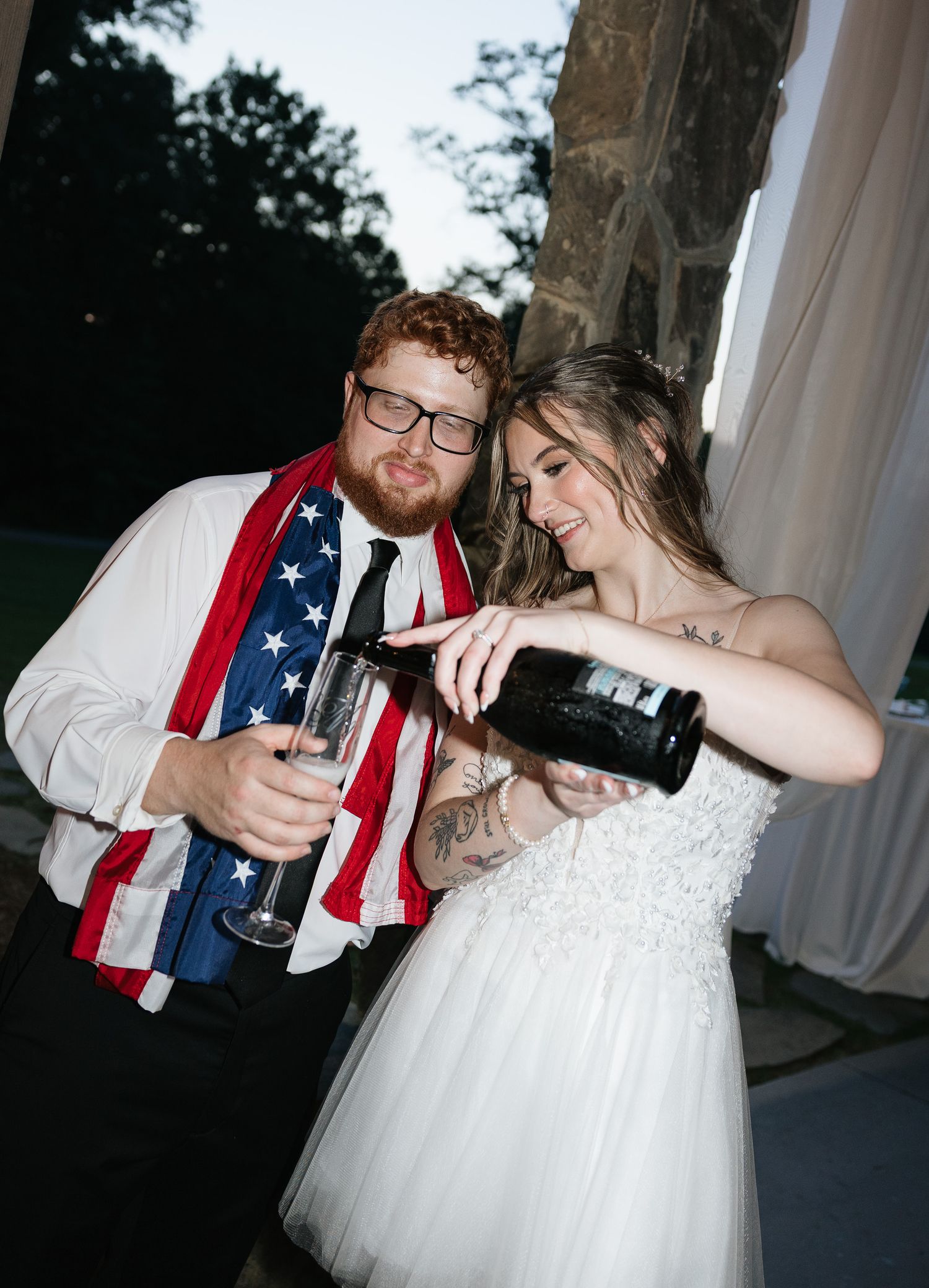 Newlyweds pouring champagne at wedding reception, groom wearing American flag and glasses, bride in white lace gown.