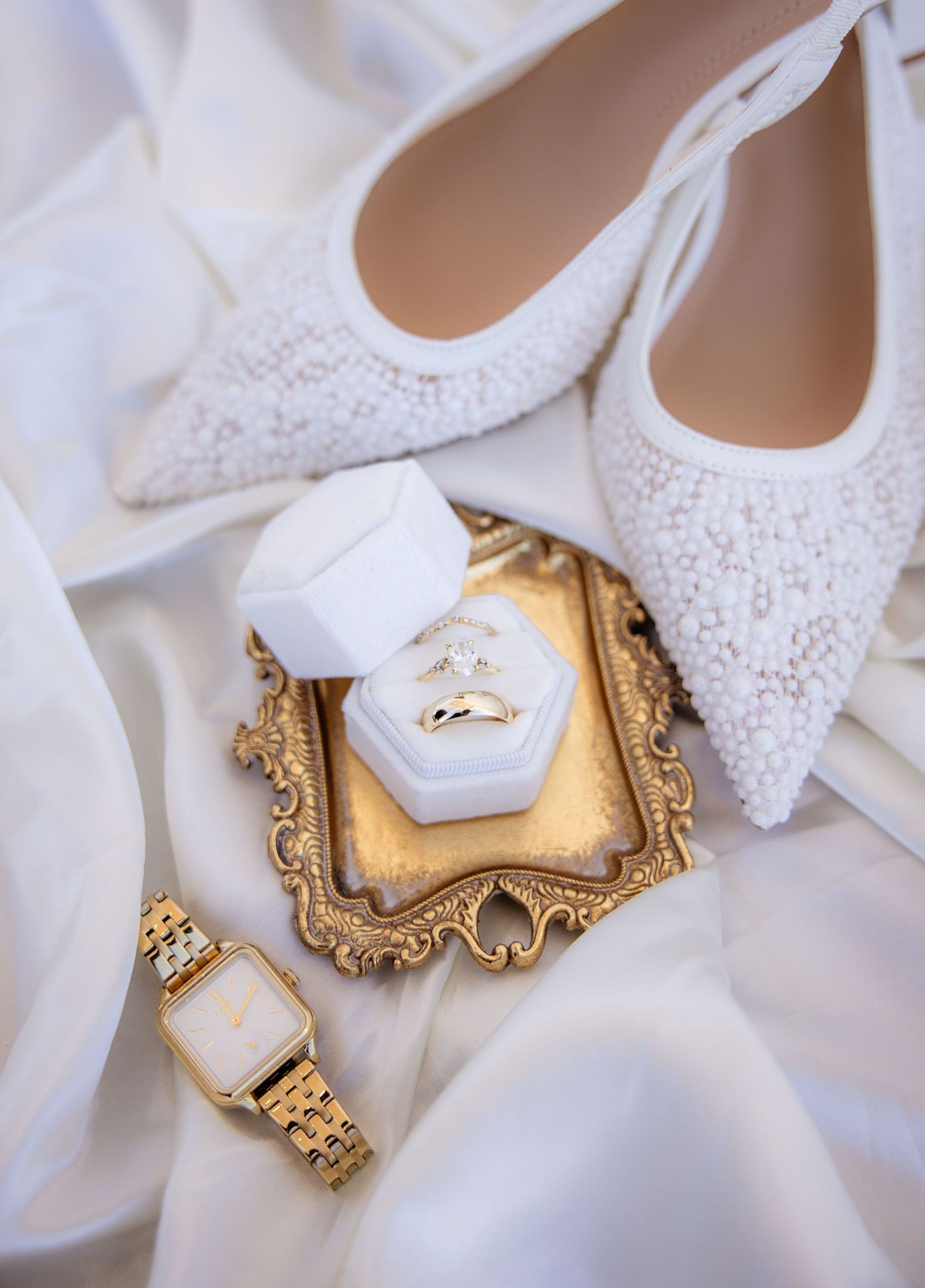 Wedding rings displayed on ornate gold frames with delicate white lace bridal shoes in the background.