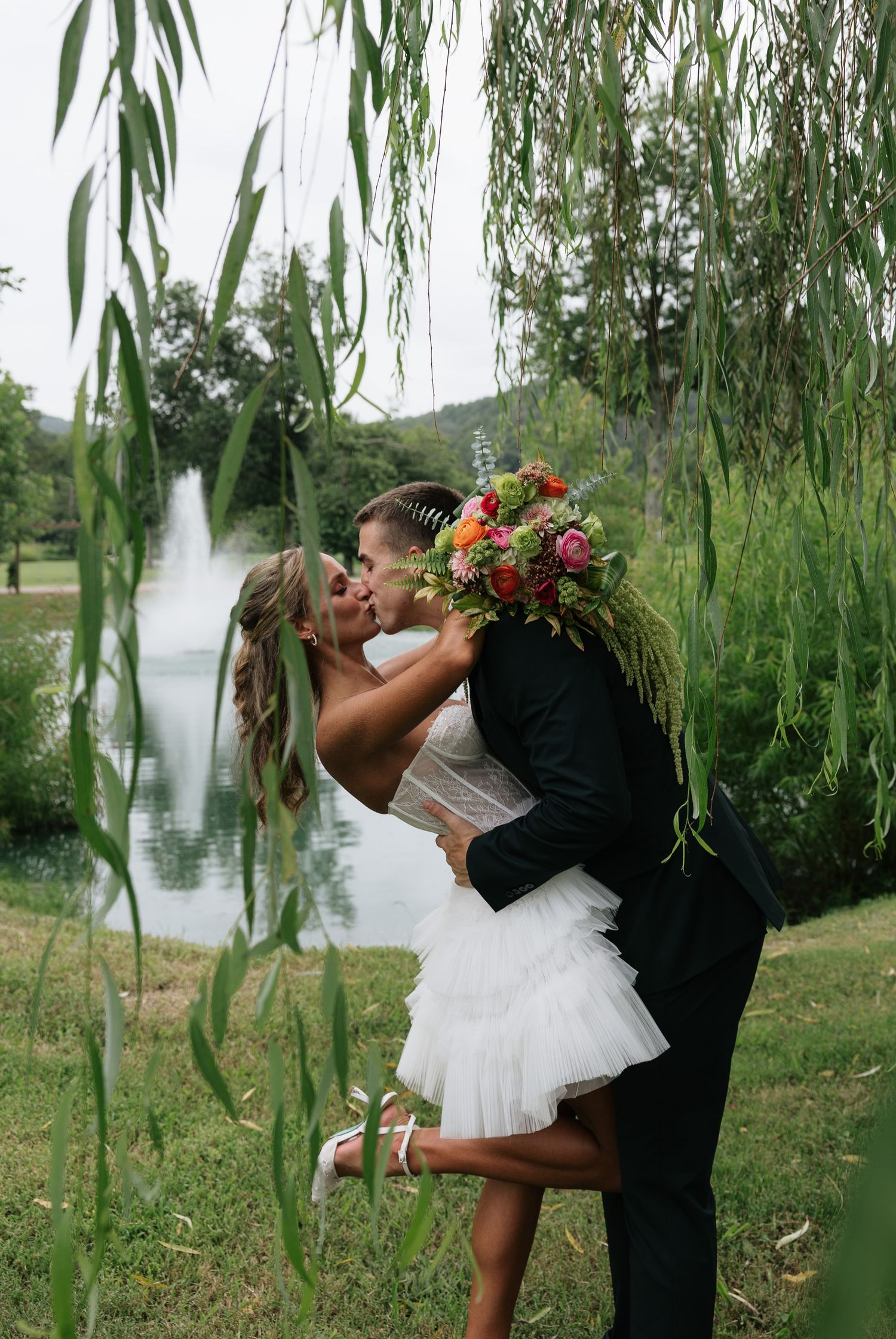 Bride and groom share romantic kiss under weeping willow tree by pond, bride holding colorful bouquet in wedding attire.
