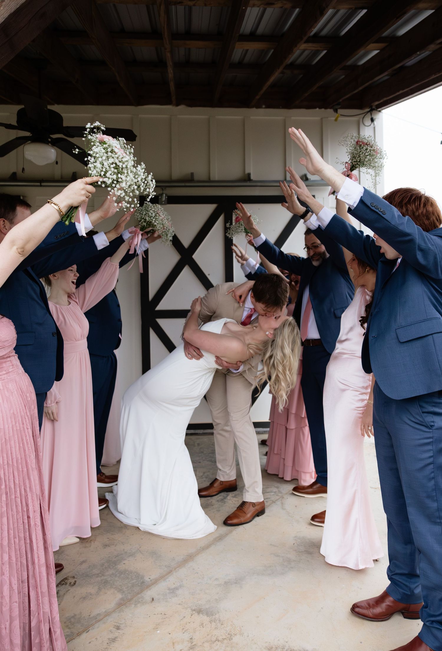 Newlyweds share a dip kiss under an arch of raised bouquets formed by wedding party in pink and navy attire.