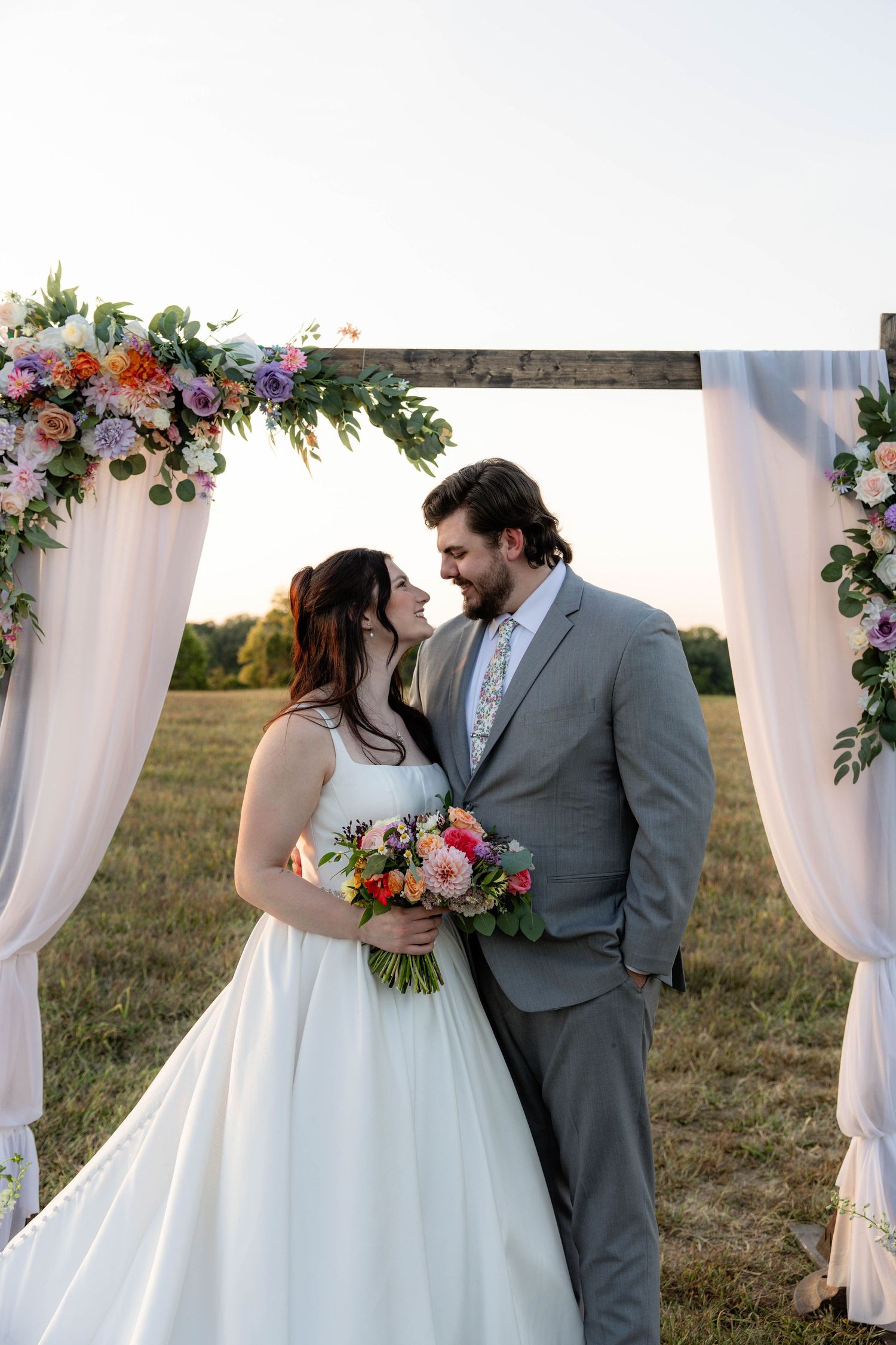 Bride and groom share a romantic moment under a floral arch with colorful bouquet at an outdoor wedding ceremony.