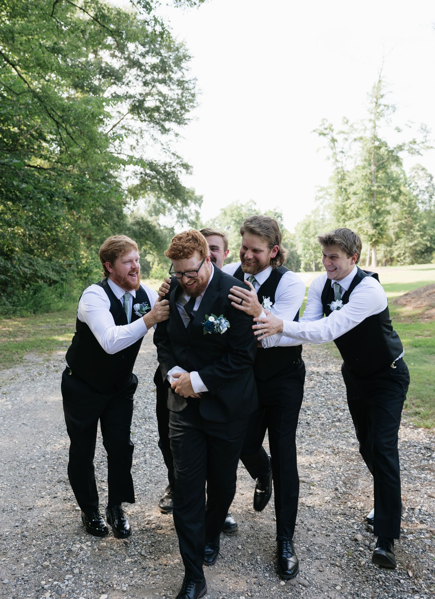 Groom and groomsmen in black vests and white shirts laugh together on gravel path at outdoor wedding.