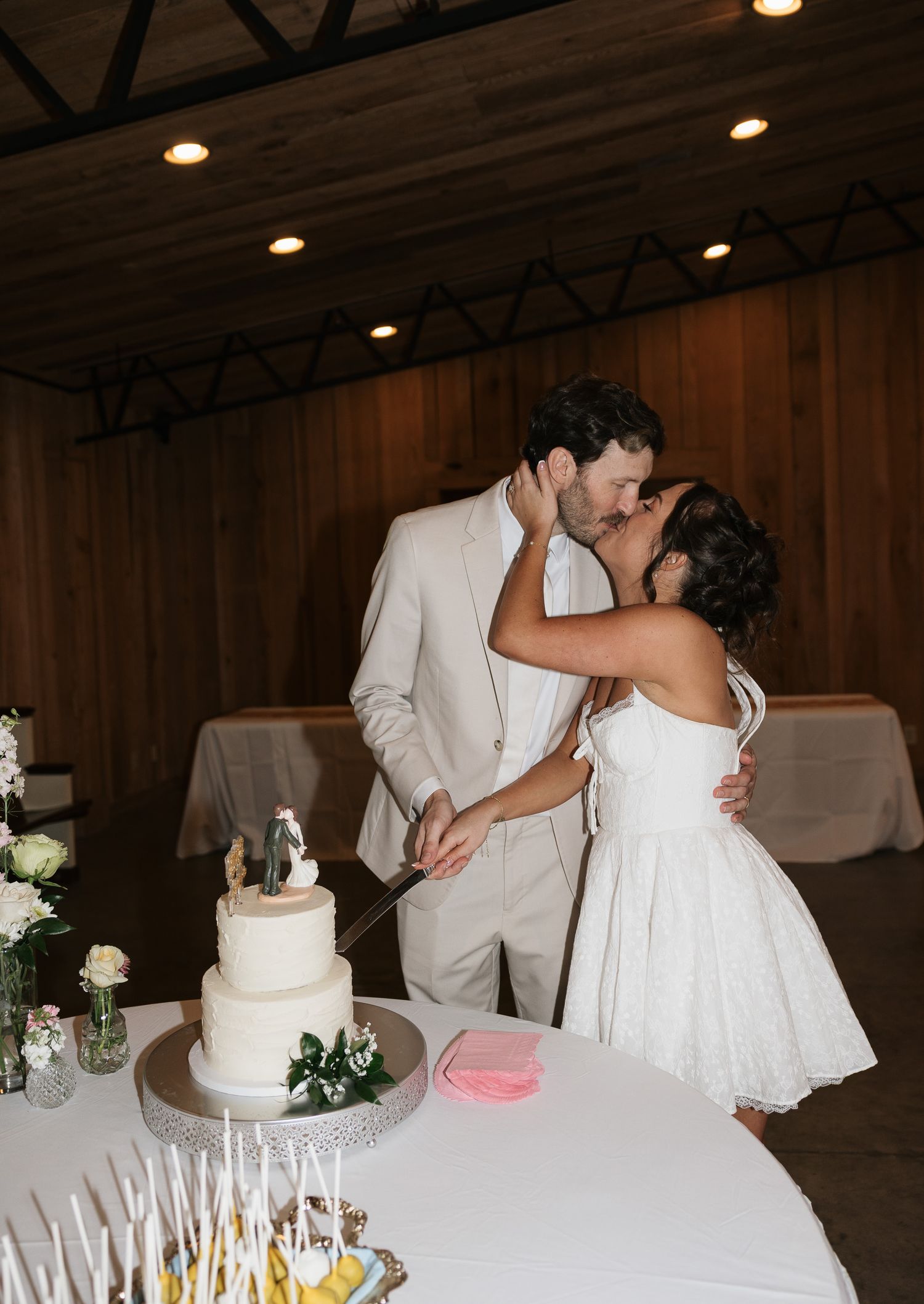 Bride and groom sharing a kiss while cutting their white wedding cake at a rustic barn reception venue.
