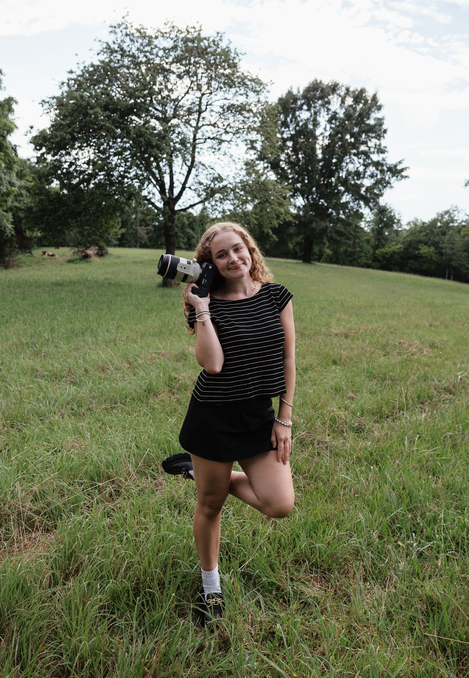 Young woman holding a camera in a lush green field, wearing a striped top and black skirt.