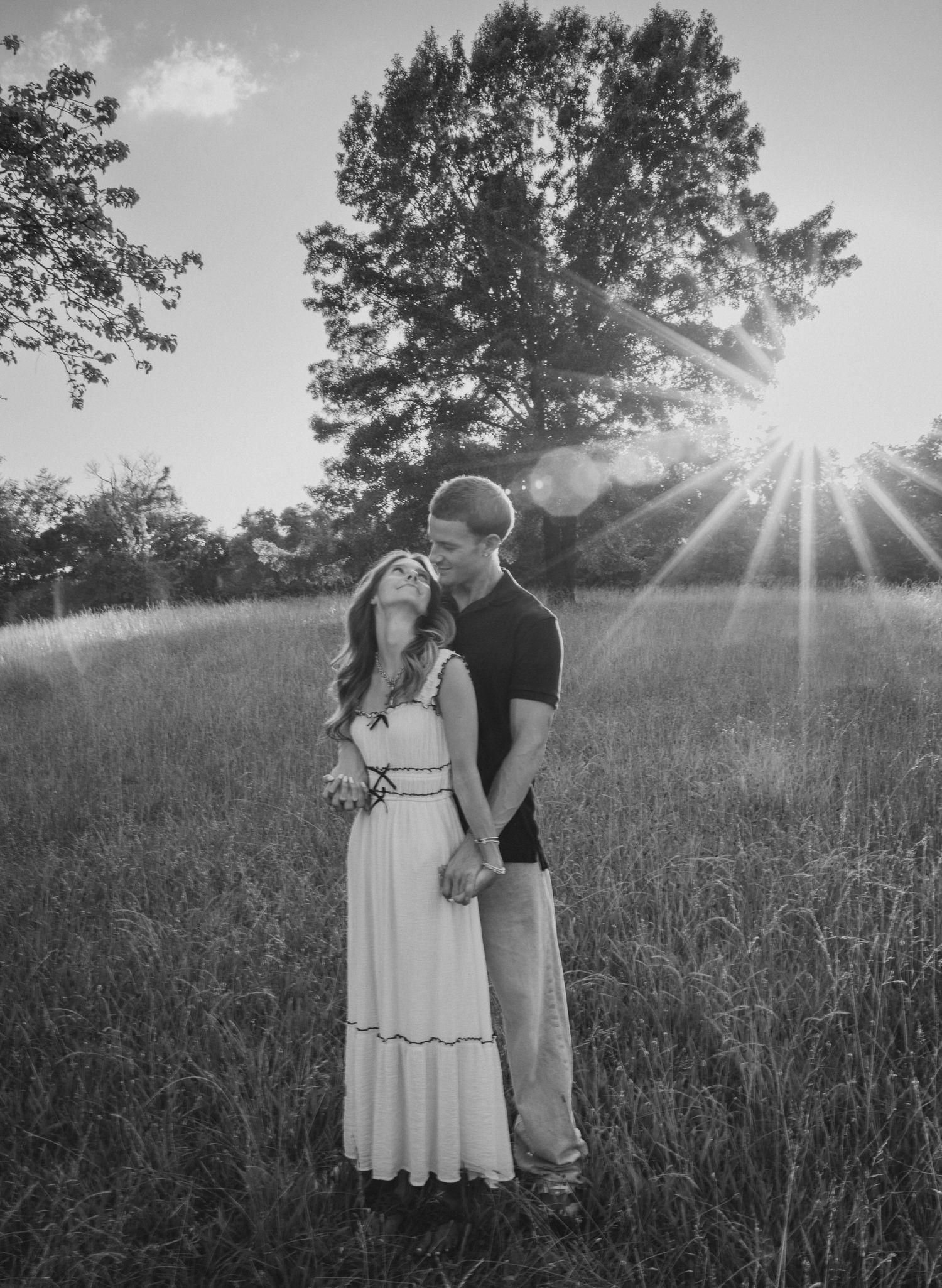 Black and white photo of a couple holding hands in a sunlit field with trees in the background.
