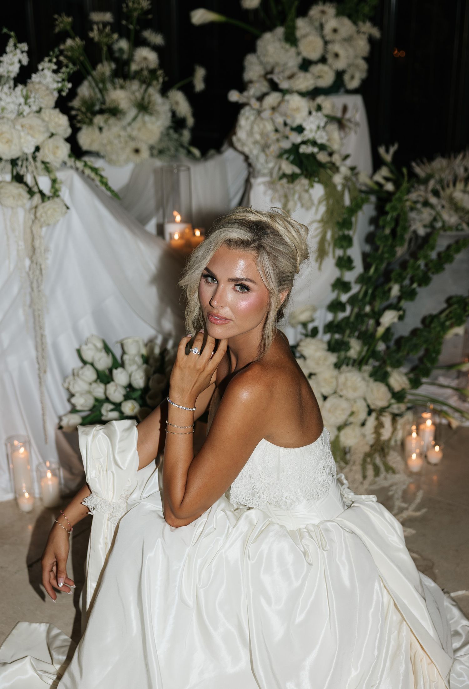 Bride in a white strapless ball gown seated among white floral arrangements and candles, looking over her shoulder.