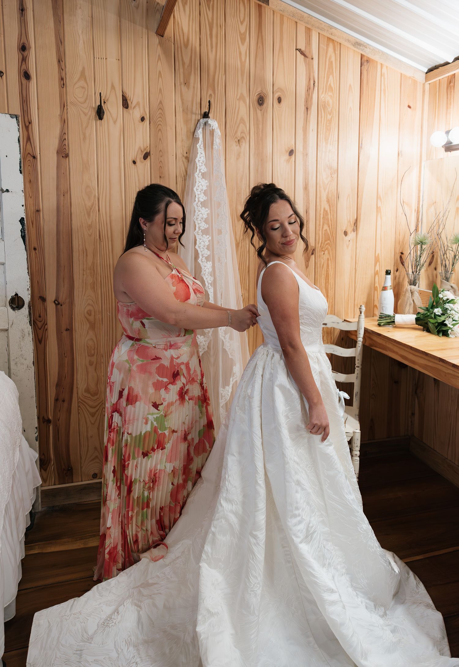 A bride in a white gown and veil being assisted by a woman in a floral dress inside a rustic wooden bridal suite.